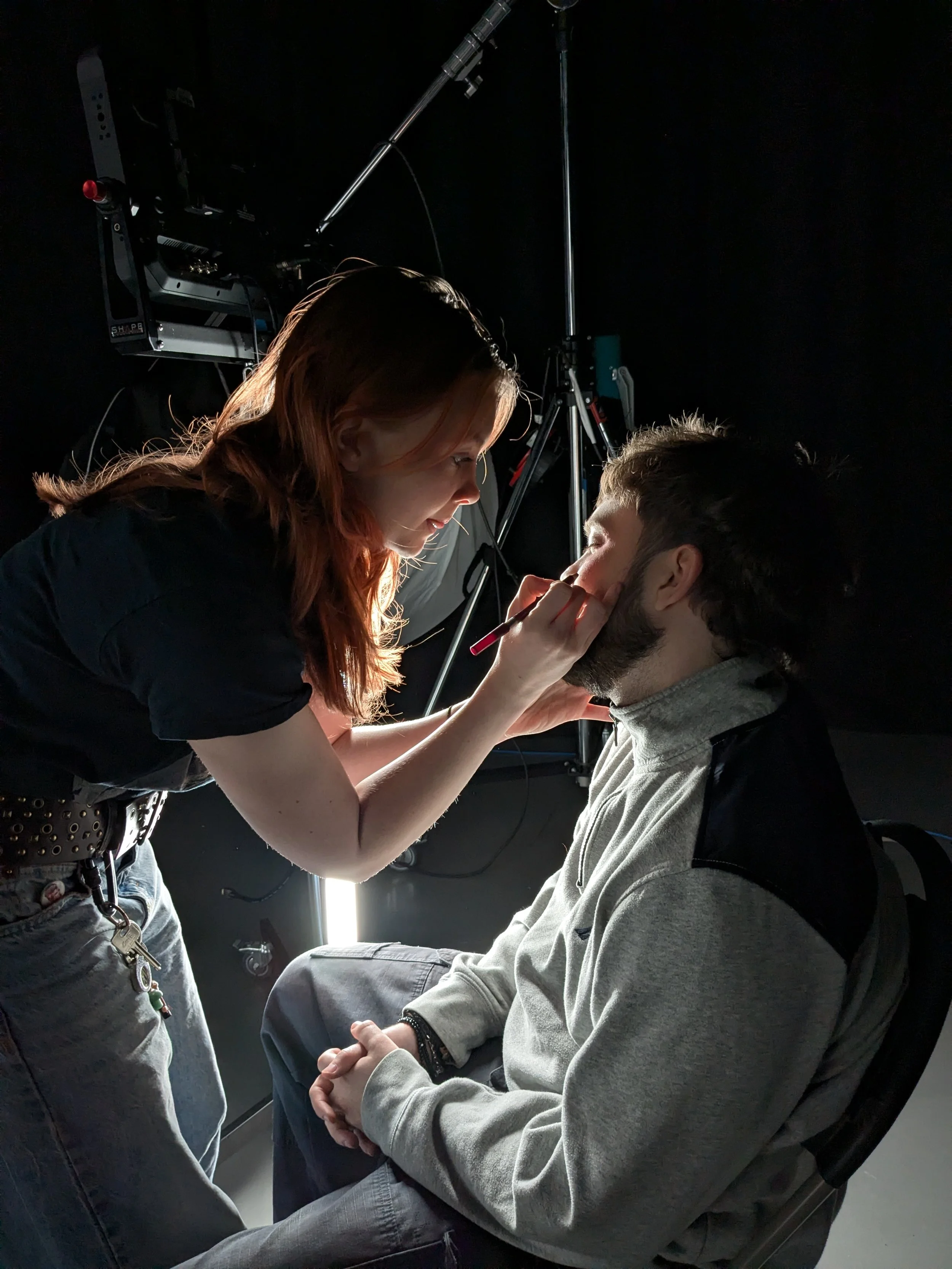 A makeup artist applying makeup to a man with long hair, seated in a makeup chair, in a professional studio setting.