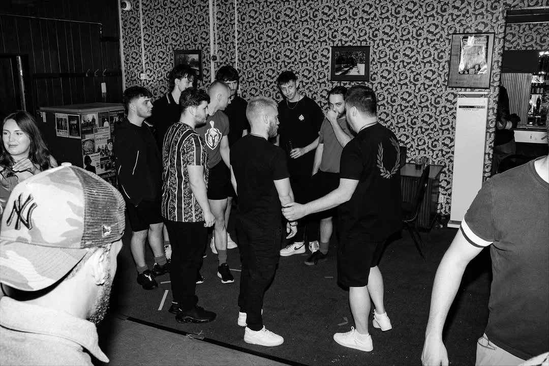 Group of young people gathered in a room, some holding hands and appearing to be engaged in a group activity, with patterned wallpaper and framed pictures on the walls.