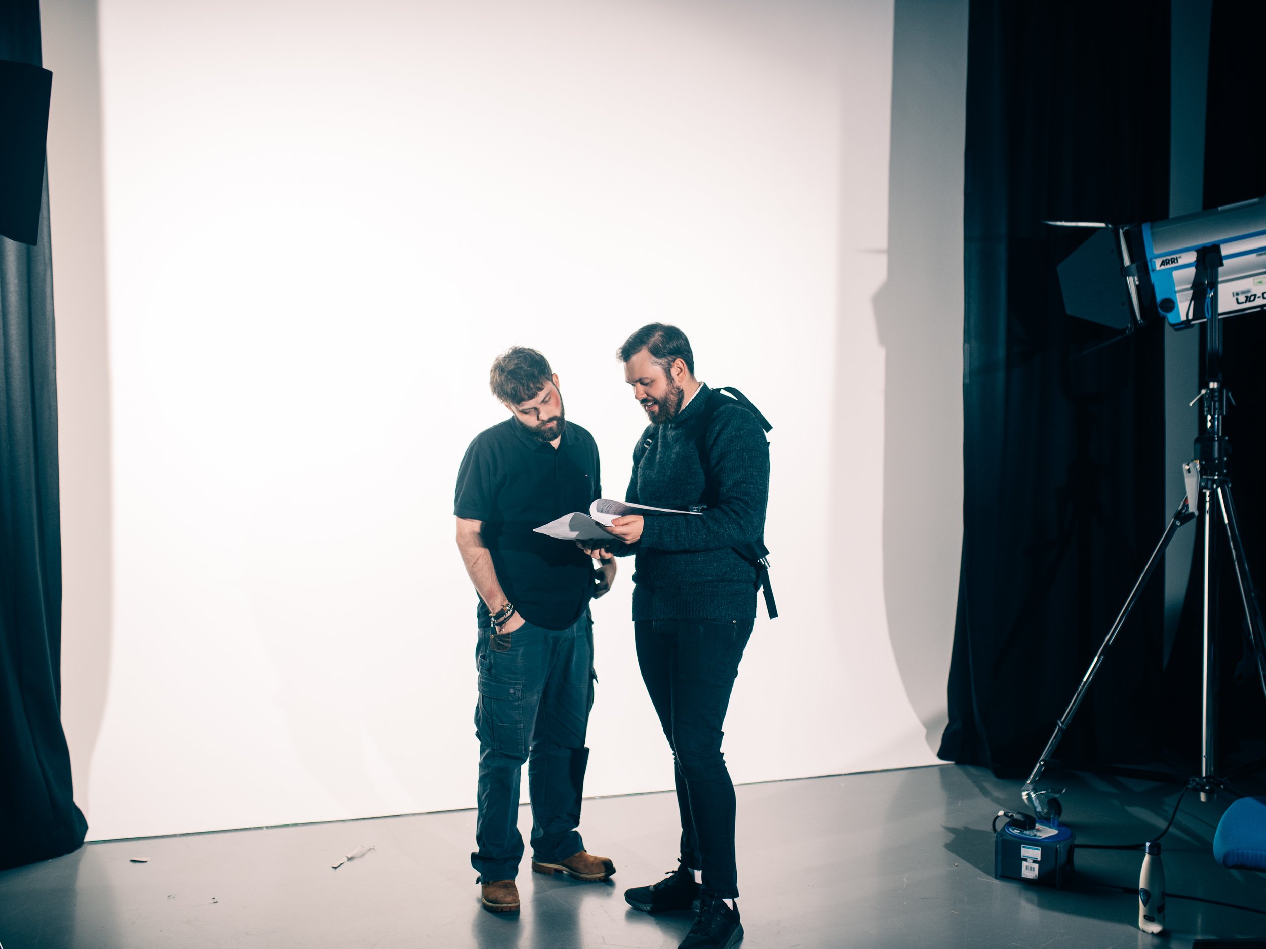 Two men standing in a photography studio, discussing something in a script or a book, with photography equipment visible to the right.