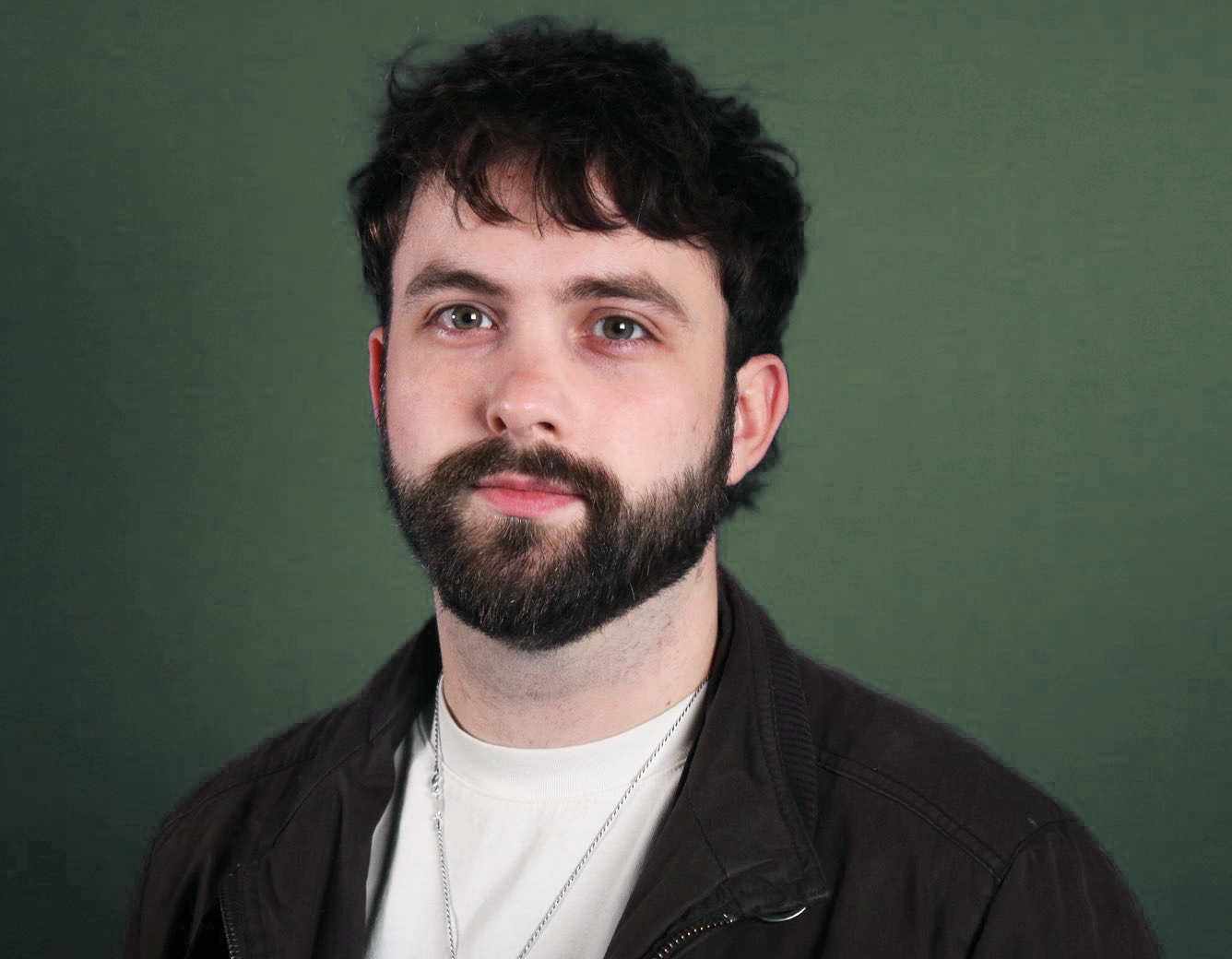 A close-up portrait of a young man with dark, curly hair and a full beard, wearing a black jacket and a white shirt, against a green background.