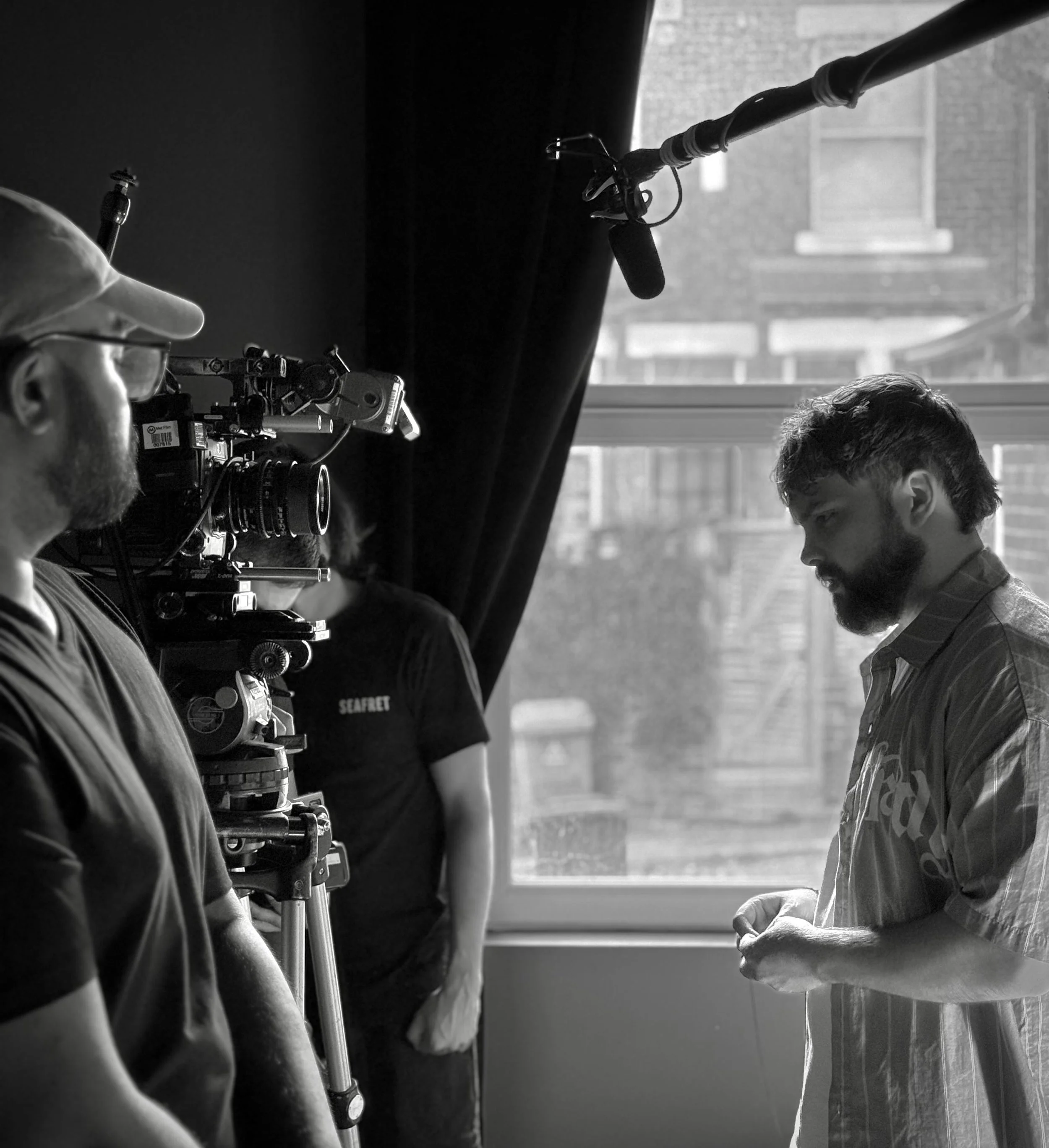 Filming scene with a man in a casual shirt looking down, and a camera operator aiming a camera at him in front of a window with buildings outside.