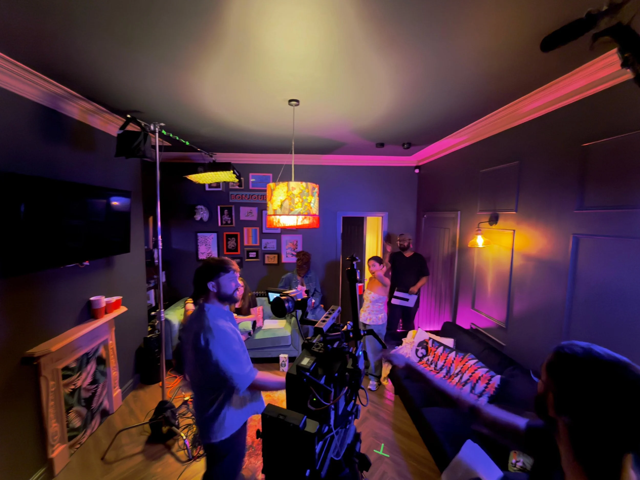 People on a film or photo shoot set in a dimly lit living room with colorful lighting, cameras, and crew members preparing for filming.