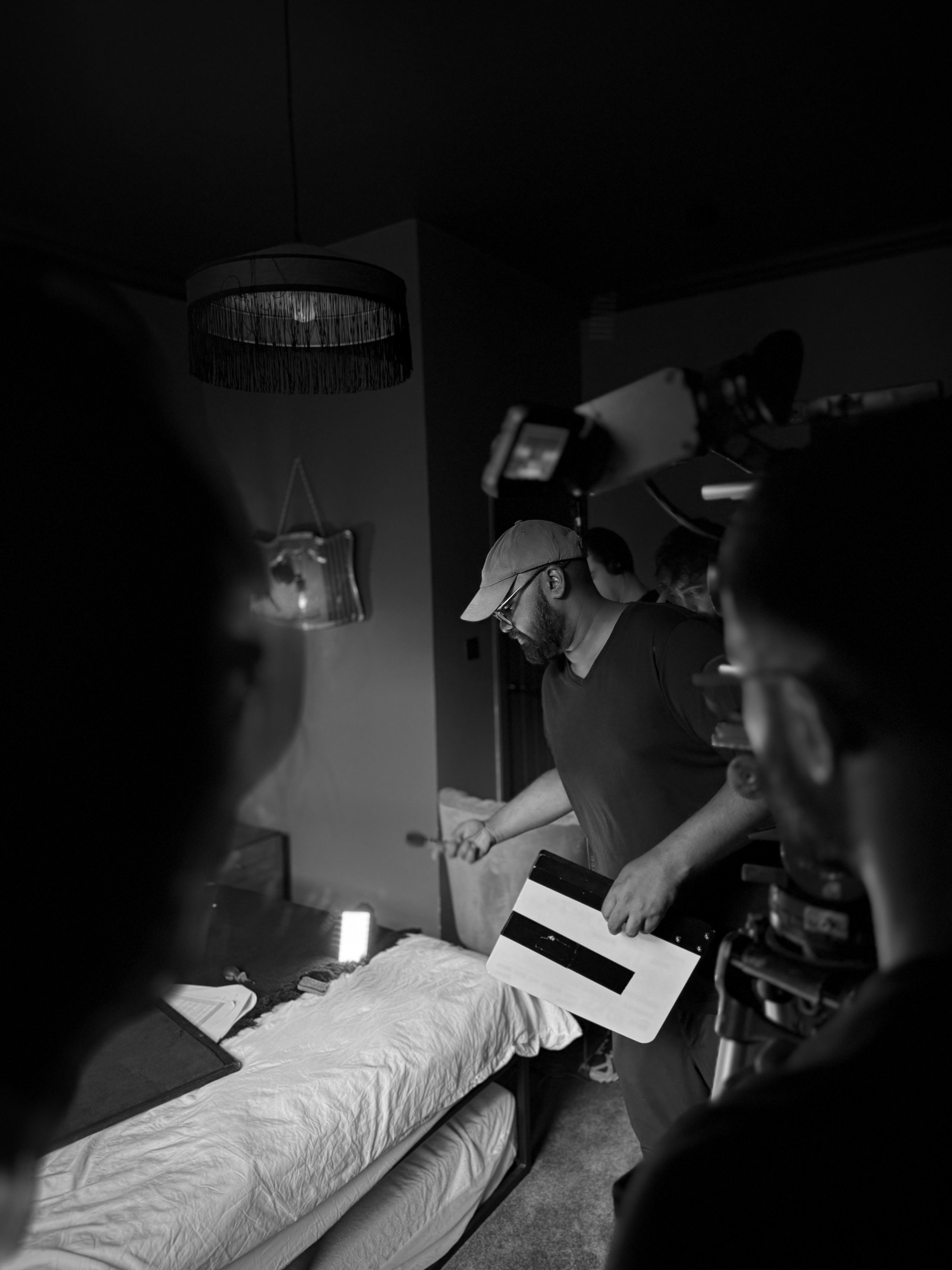 A man in a cap and glasses holding a clapperboard on a film set, with crew members around him, filming a scene in a bedroom.