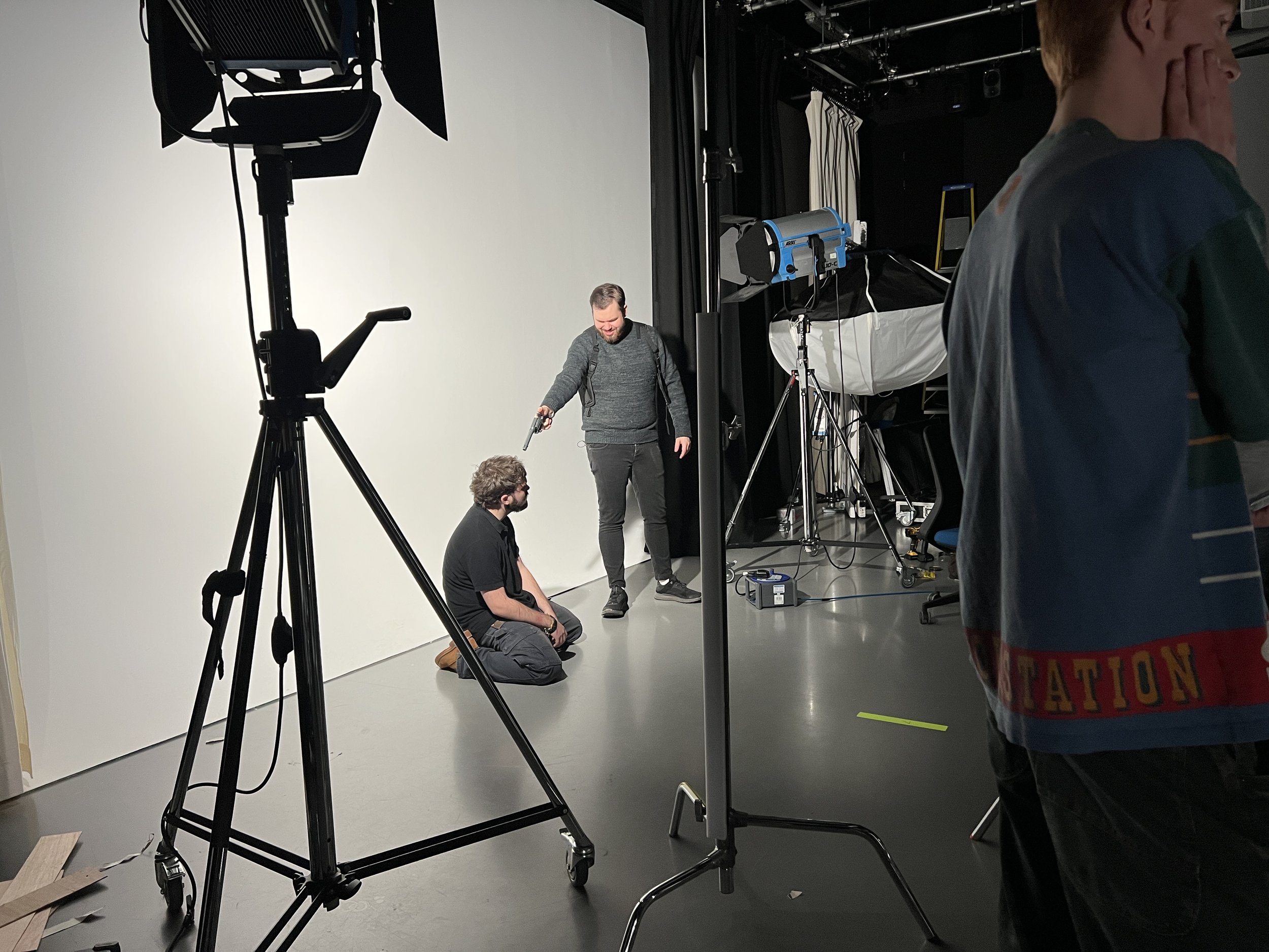 People on a film or photo set, with one person sitting on the floor and another standing, surrounded by studio lighting and equipment.