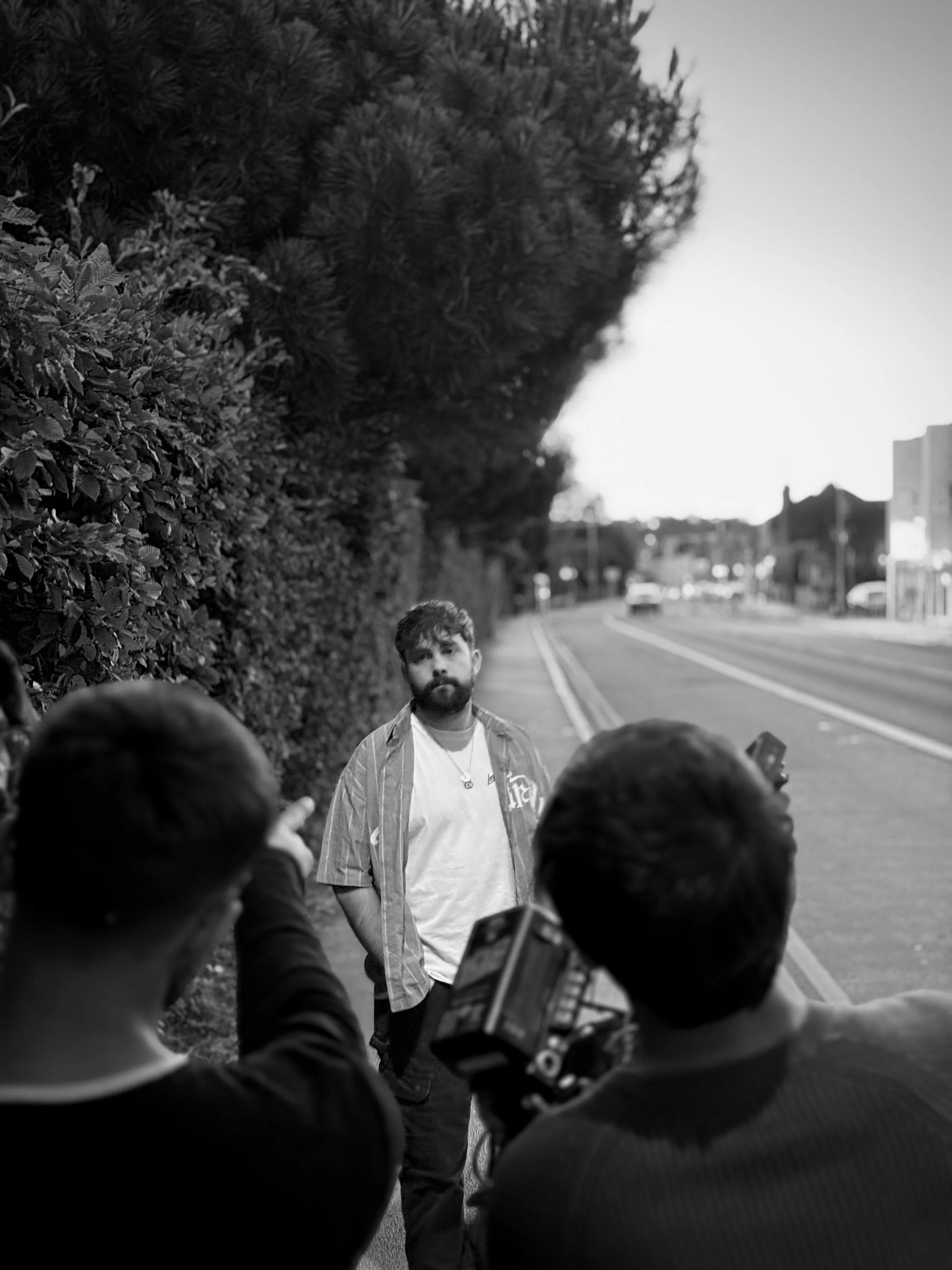 A man with curly hair and a beard standing on a sidewalk with trees and a road behind him, while two people are filming or taking pictures of him.