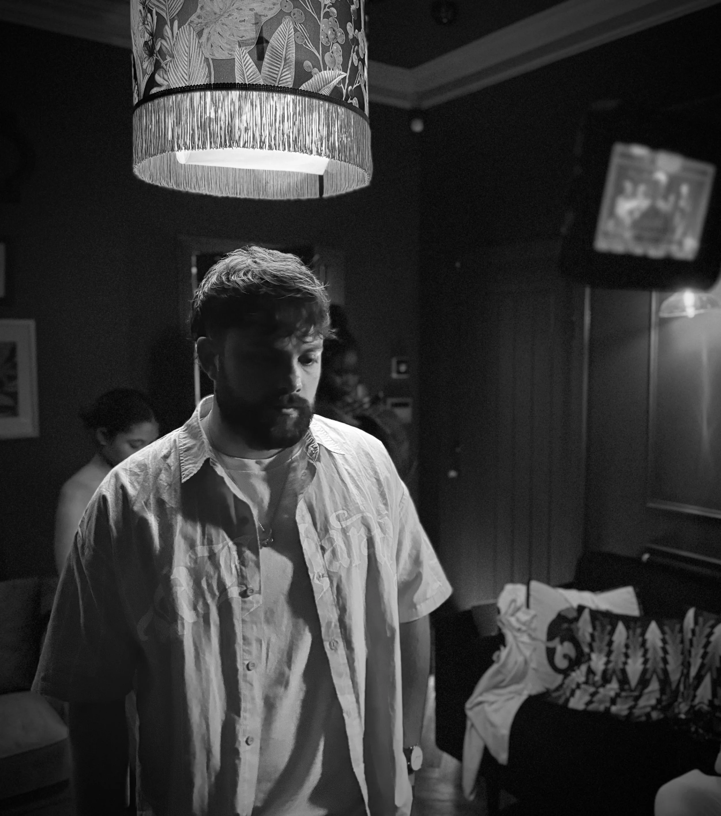 A man with a beard and wearing a light-colored shirt stands in a dimly lit room, with a patterned lampshade hanging above him. In the background, there's a woman and a TV mounted on the wall.