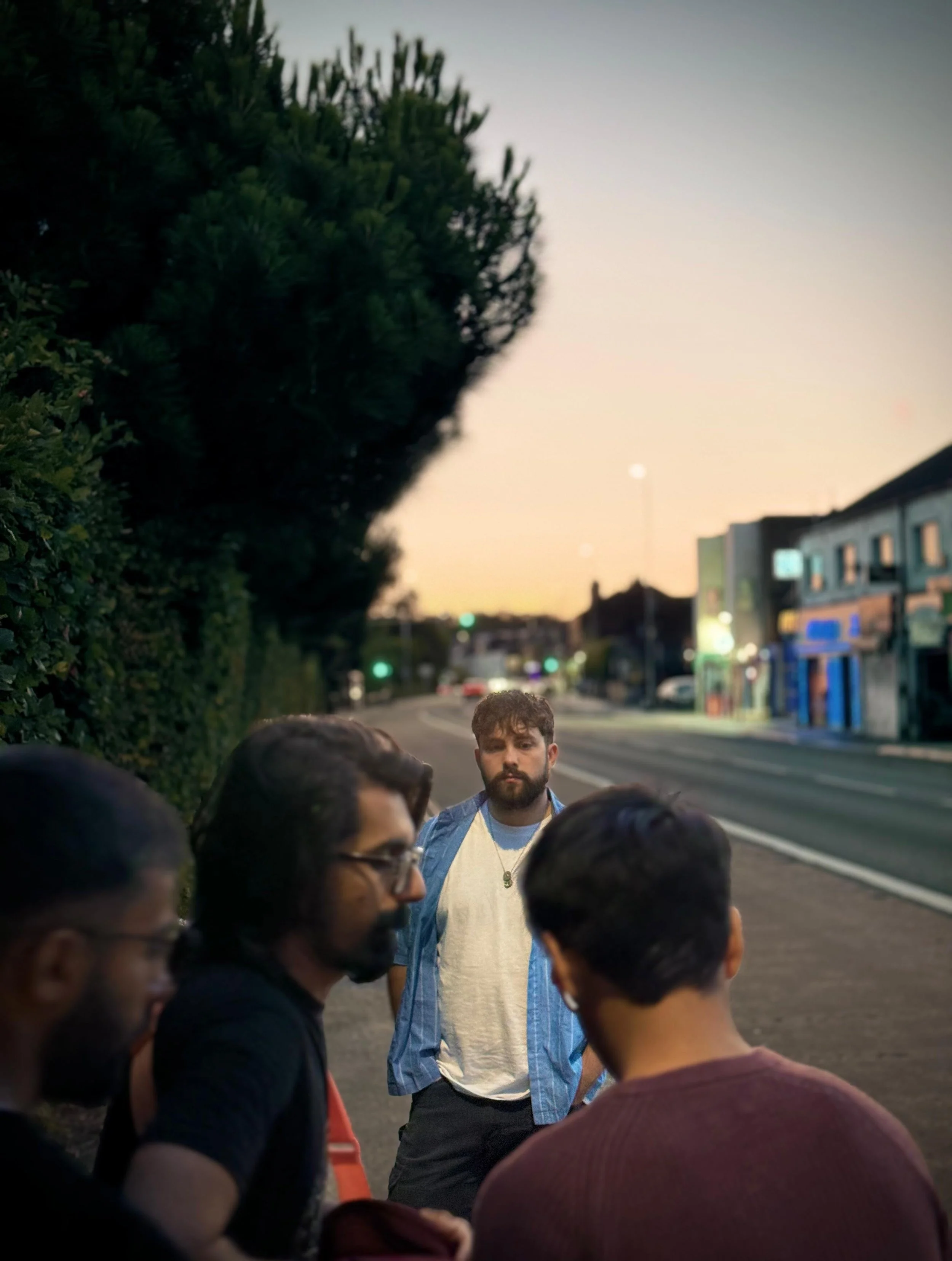 Four men standing and talking on a city sidewalk during dusk, with a large tree, commercial buildings, and streetlights in the background.