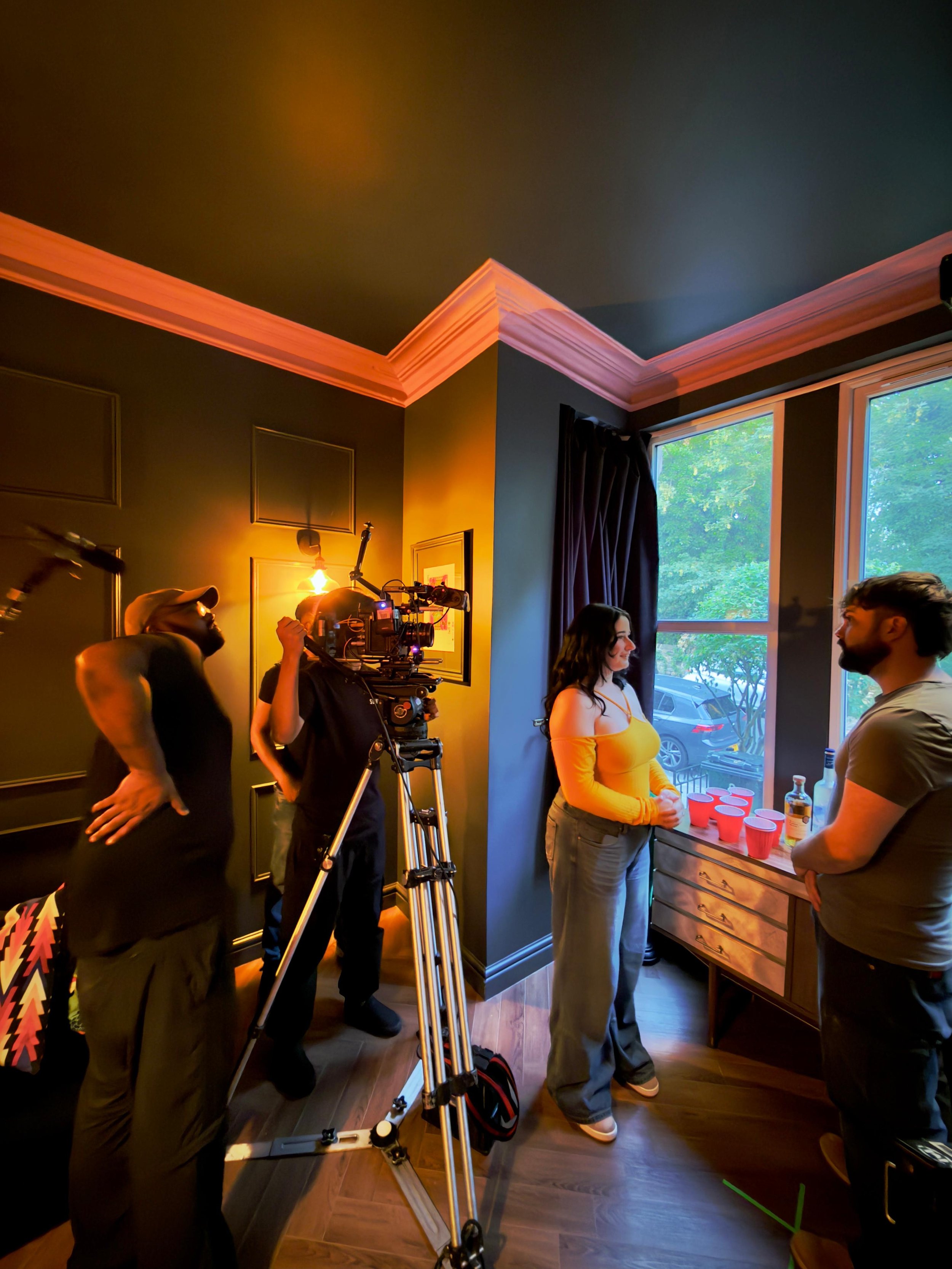 Behind the scenes of a video shoot or interview in a warmly lit room with dark green walls, a large window showing green trees outside, two people talking near the window, and film crew members operating a camera.