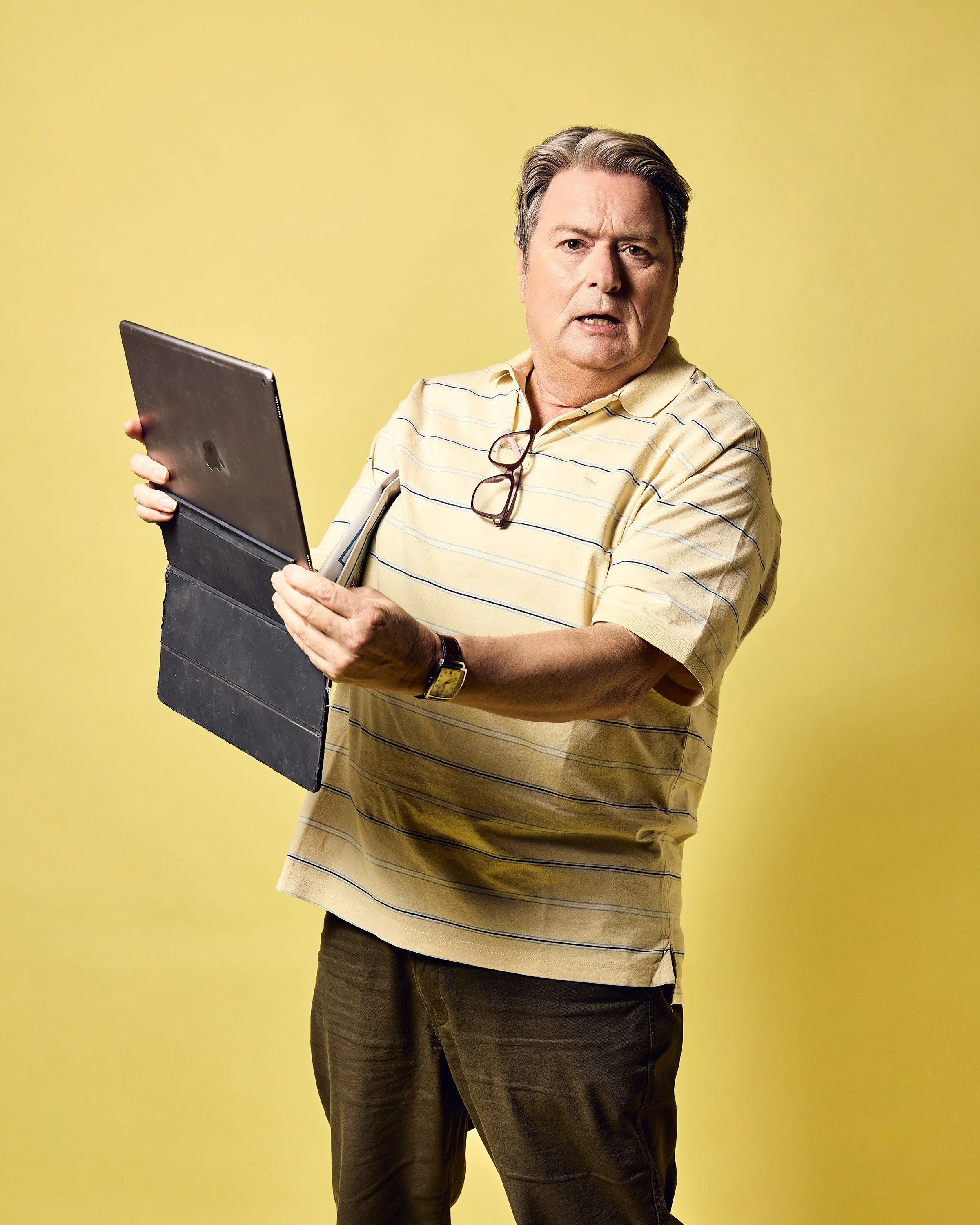 A middle-aged man with gray hair wearing a beige striped polo shirt, holding a laptop and folder, with a surprised or confused expression on his face, standing against a yellow background.