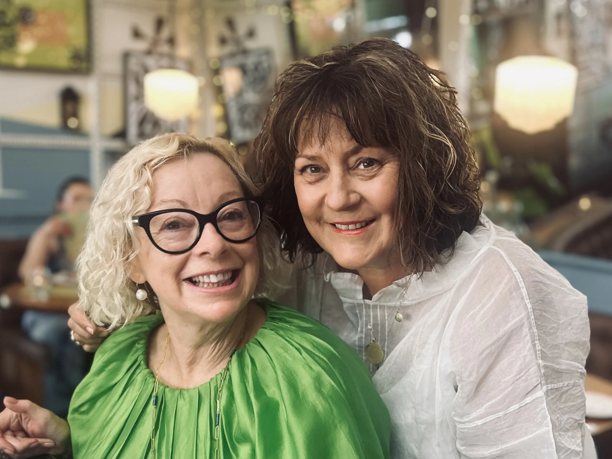 Two women smiling, one in a green top with glasses and the other in a white top, in a restaurant with warm lighting and decorative mirrors in the background.