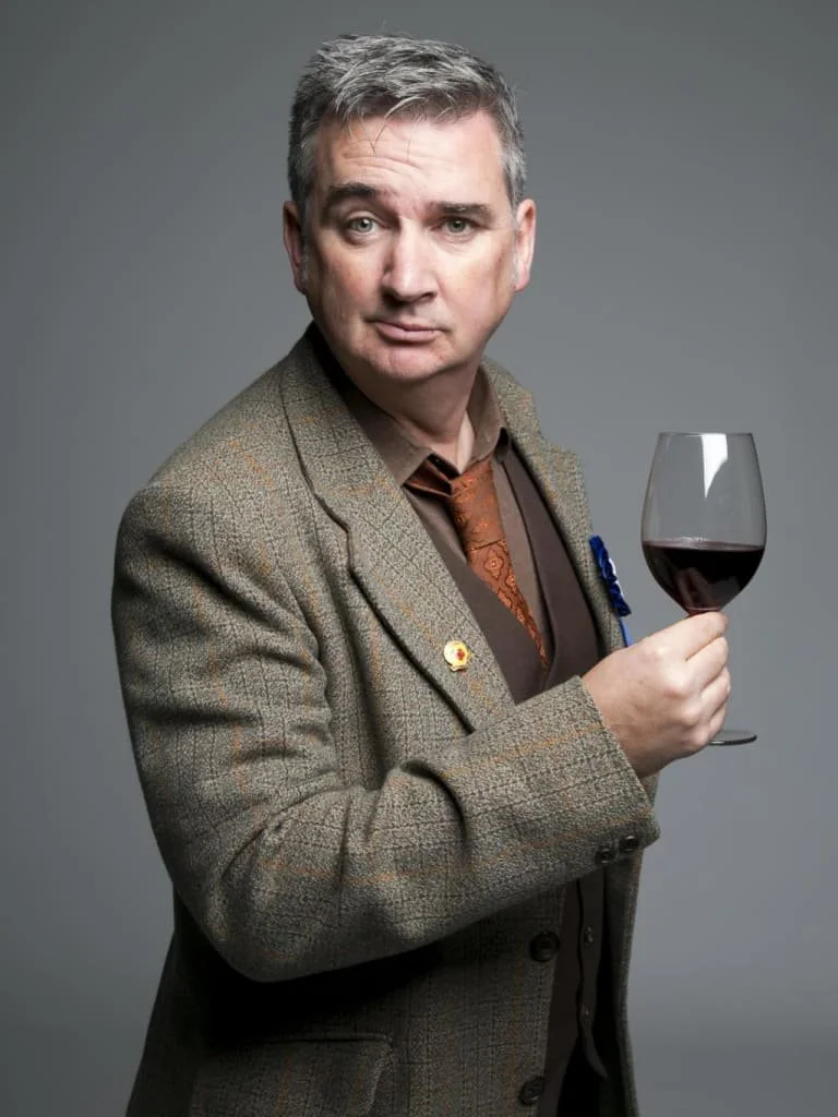 Man in a tweed jacket holding a glass of red wine, looking at the camera with a slightly raised eyebrow against a gray background.