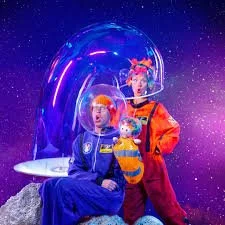 Two children dressed in colorful space-themed costumes sit on a rock in front of a futuristic spaceship with a glowing blue dome, set against a starry outer space background.
