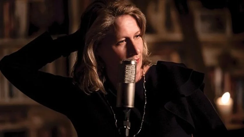 A woman with blonde hair is standing near a microphone in a room with shelves of books in the background. She has her left hand behind her head and is looking to the side, with a contemplative expression.