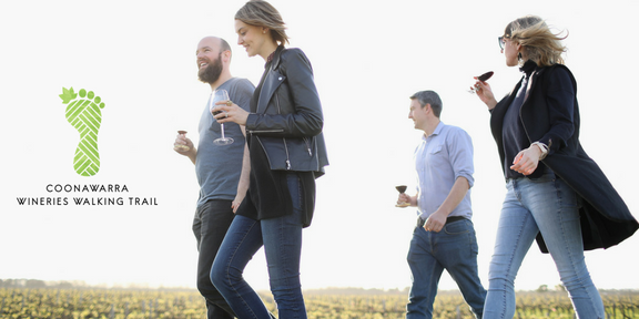 Four people walking outdoors, two women and two men, enjoying drinks and using their phones, with a logo of a green foot and the text 'Coonawarra Wineries Walking Trail'.