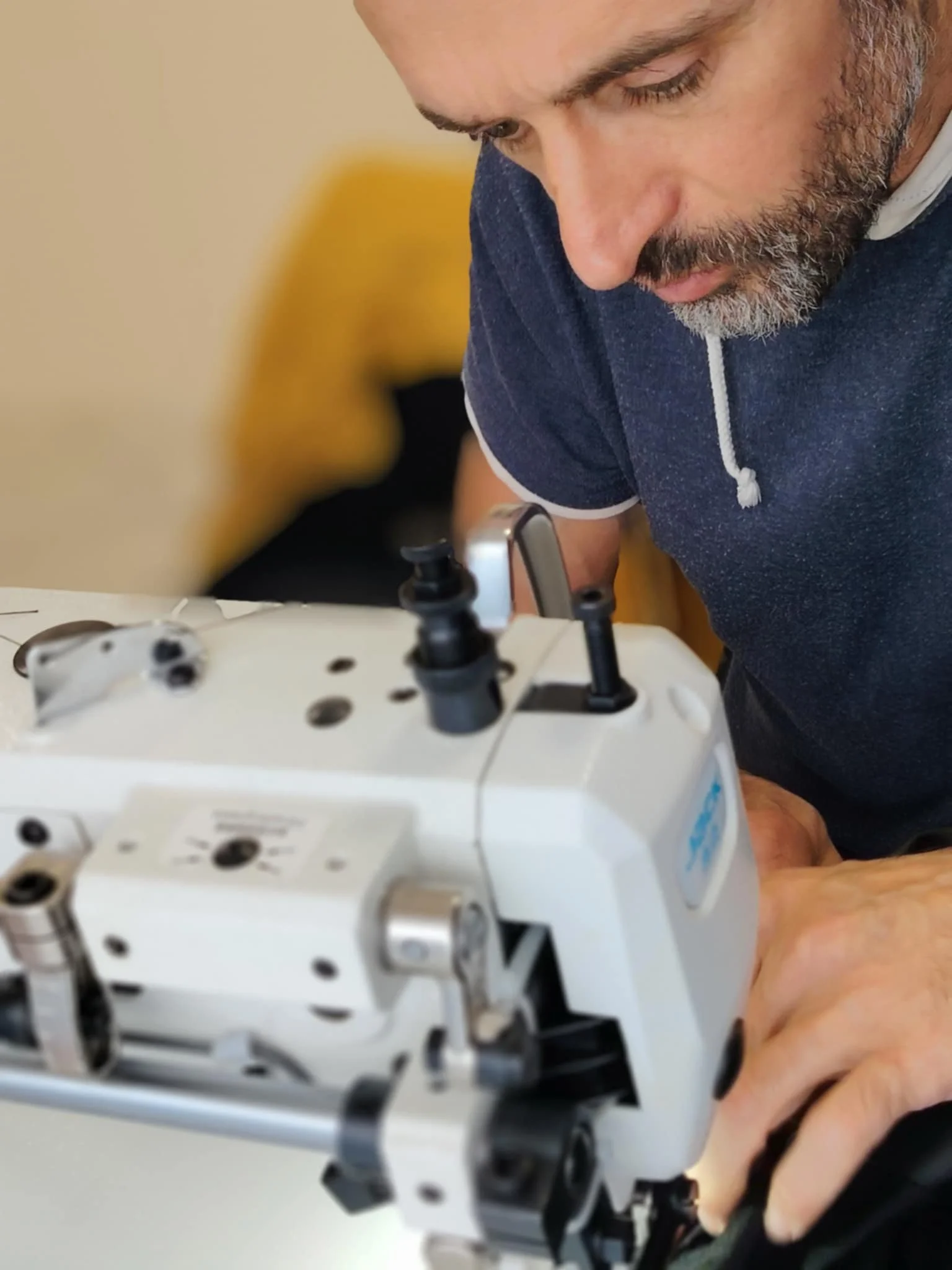 A man with a beard works intently with a piece of industrial or medical equipment, possibly a sewing machine or a piece of lab machinery, adjusting or inspecting it closely.