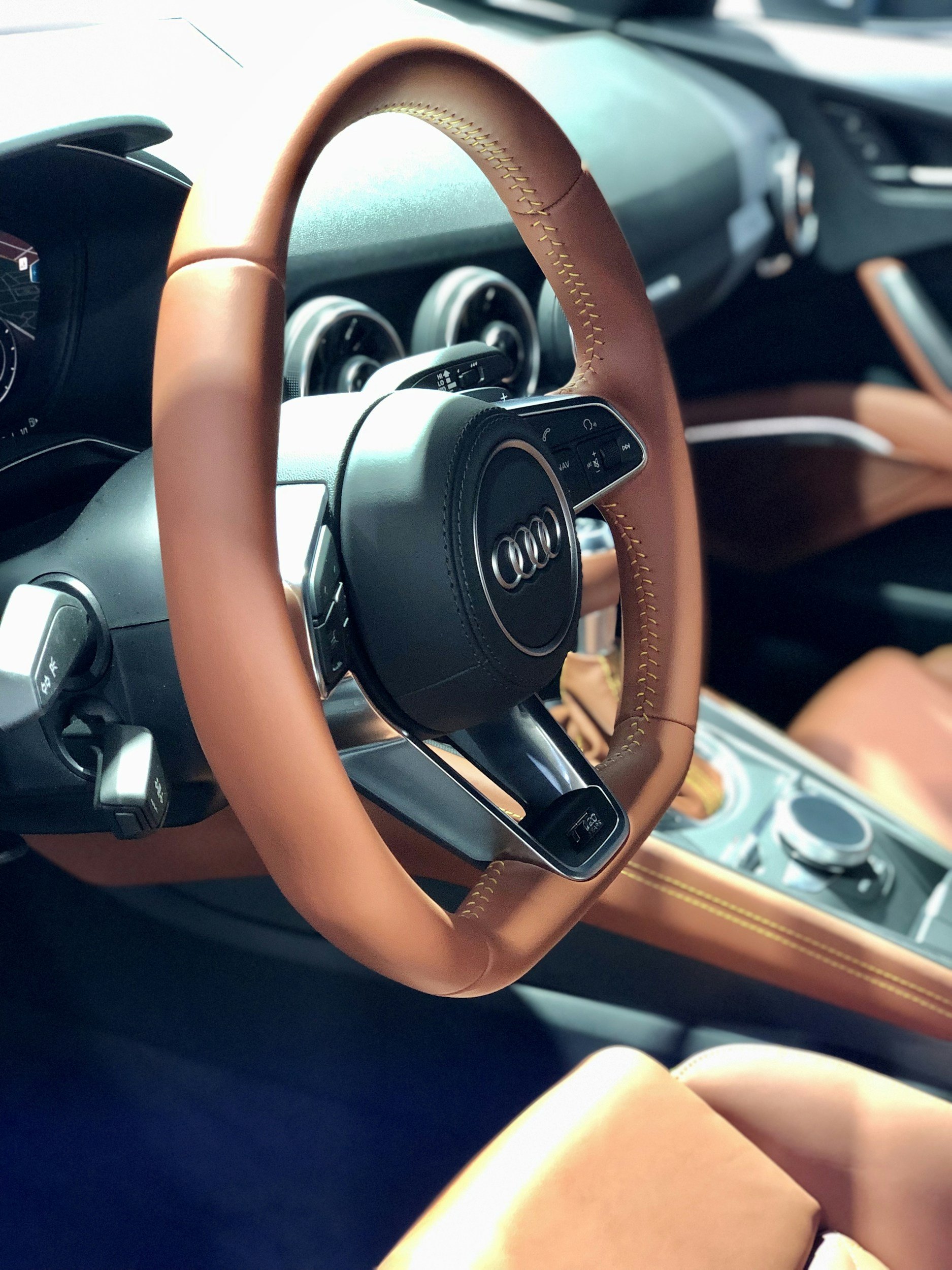 Interior of an Audi car showing a brown leather steering wheel with yellow stitching, dashboard, and center console with control knob.