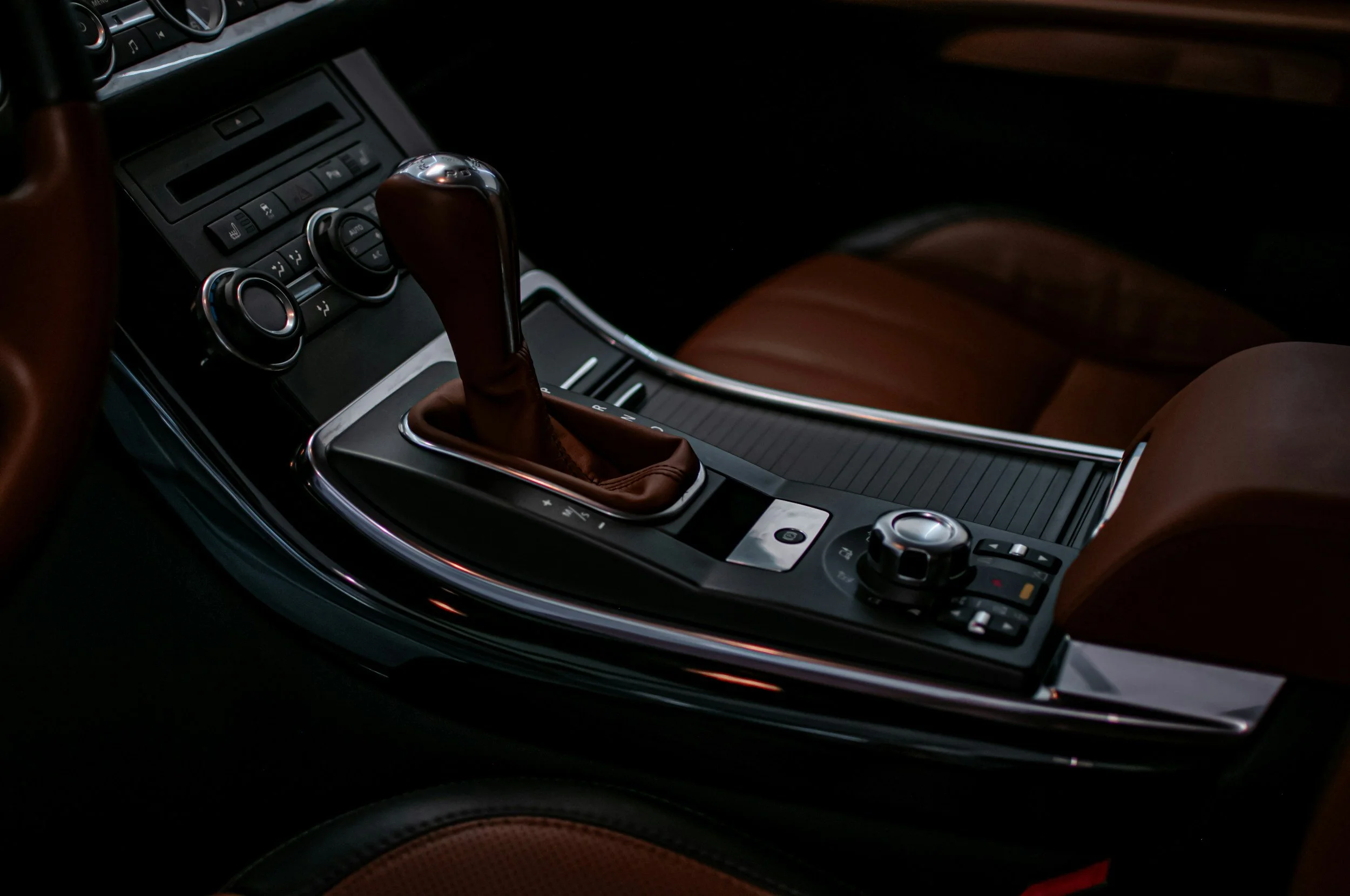 Close-up of a car's center console featuring a gear shift with a leather cover, control buttons, and a rotary knob, with brown leather seats visible.