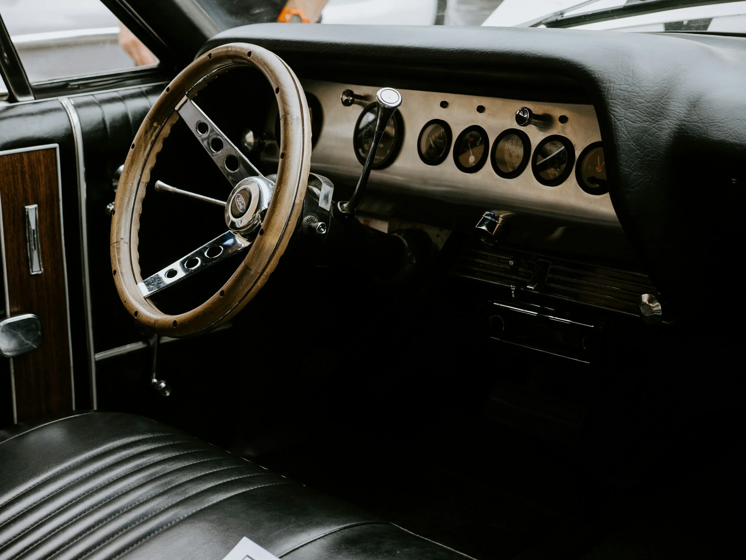 Interior of a vintage car showing a wood-rimmed steering wheel, dashboard with analog gauges, and black leather seats.