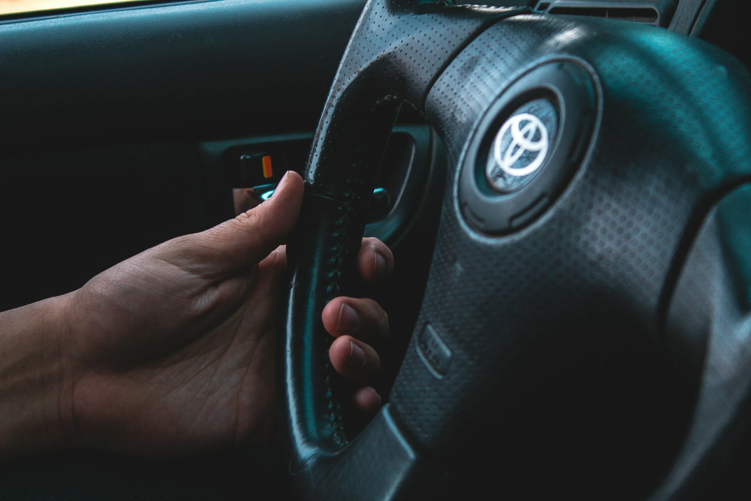 Hand gripping a car steering wheel inside vehicle, with Toyota logo on the wheel.