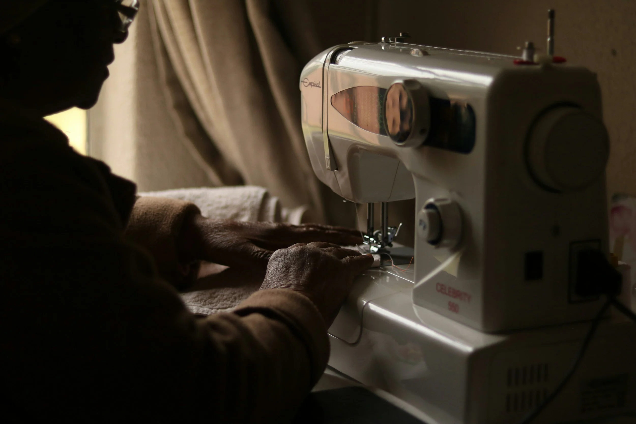 An elderly person sewing using a white sewing machine in a dimly lit room.