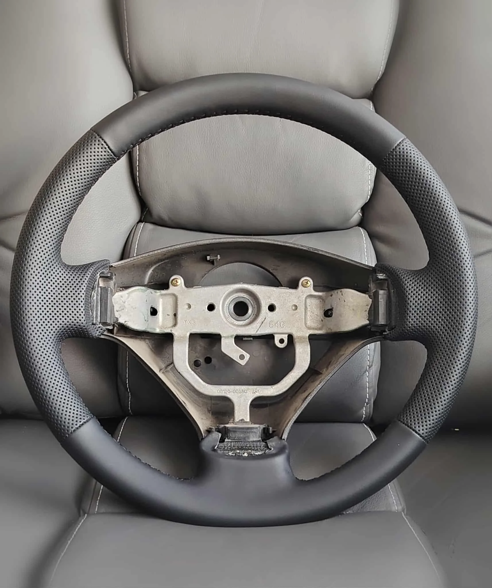 A disassembled car steering wheel with a leather grip resting on a gray leather car seat.