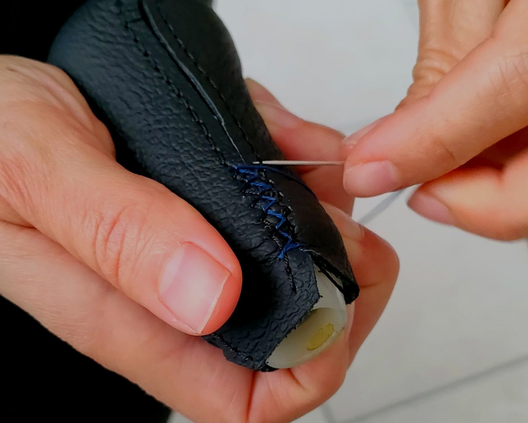 Hands stitching black leather on a shoe with a needle and blue thread.