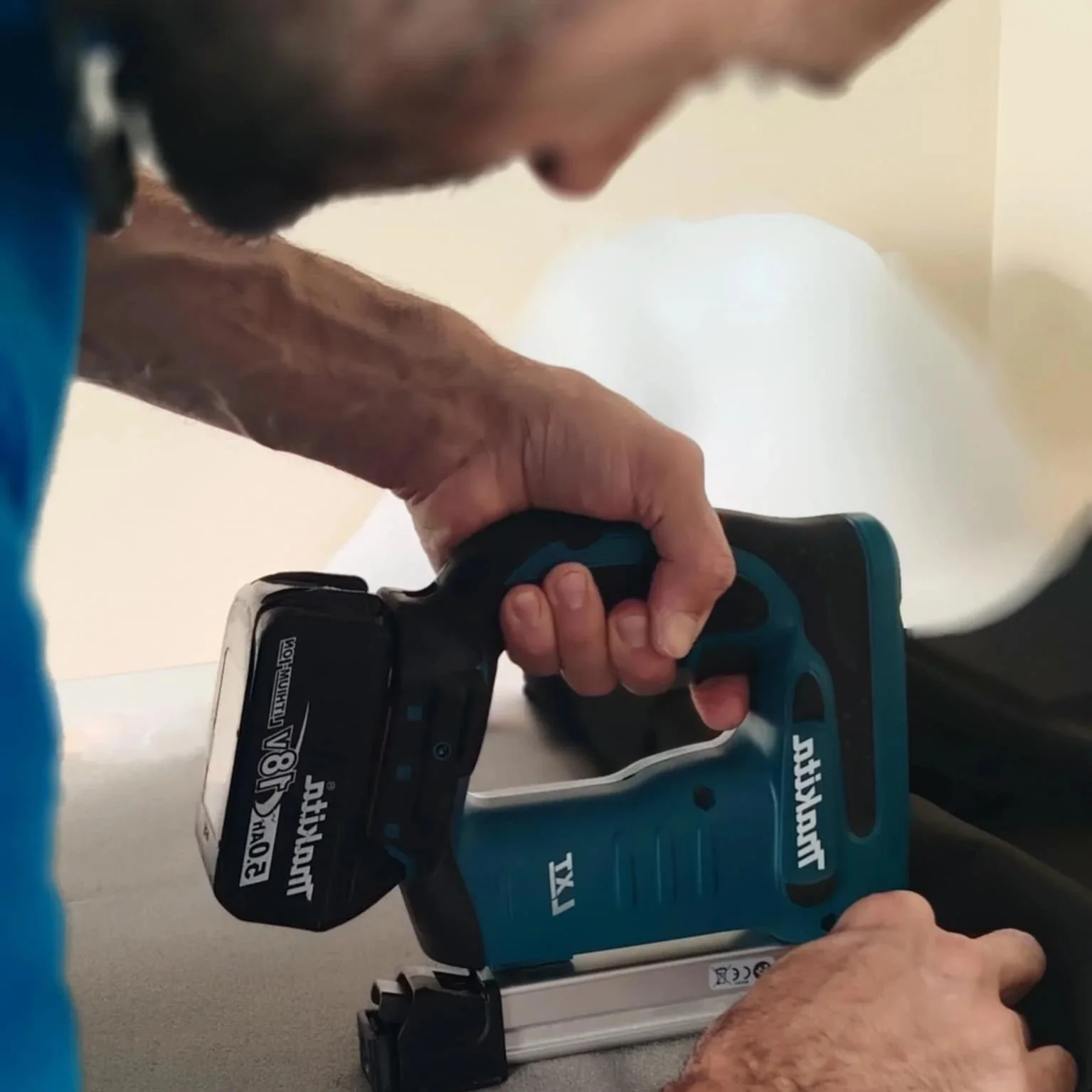 A person using a Makita cordless nail gun, pressing the trigger with their right hand while their left hand steadies the tool on a surface, with a background of a white balloon or round object.