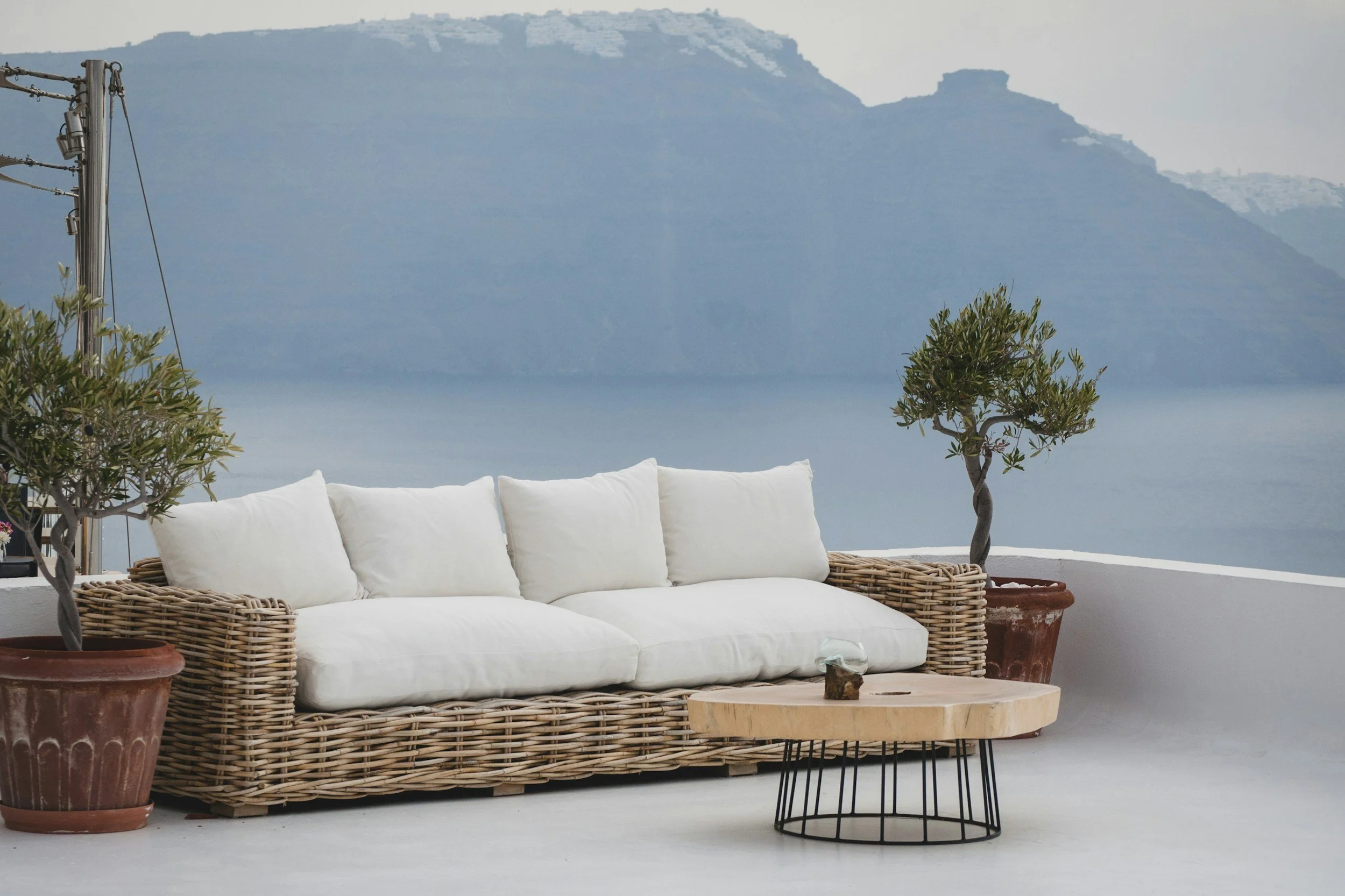 Outdoor patio with a wicker sofa with white cushions, a wooden coffee table, and potted plants, overlooking a body of water and mountains in the distance