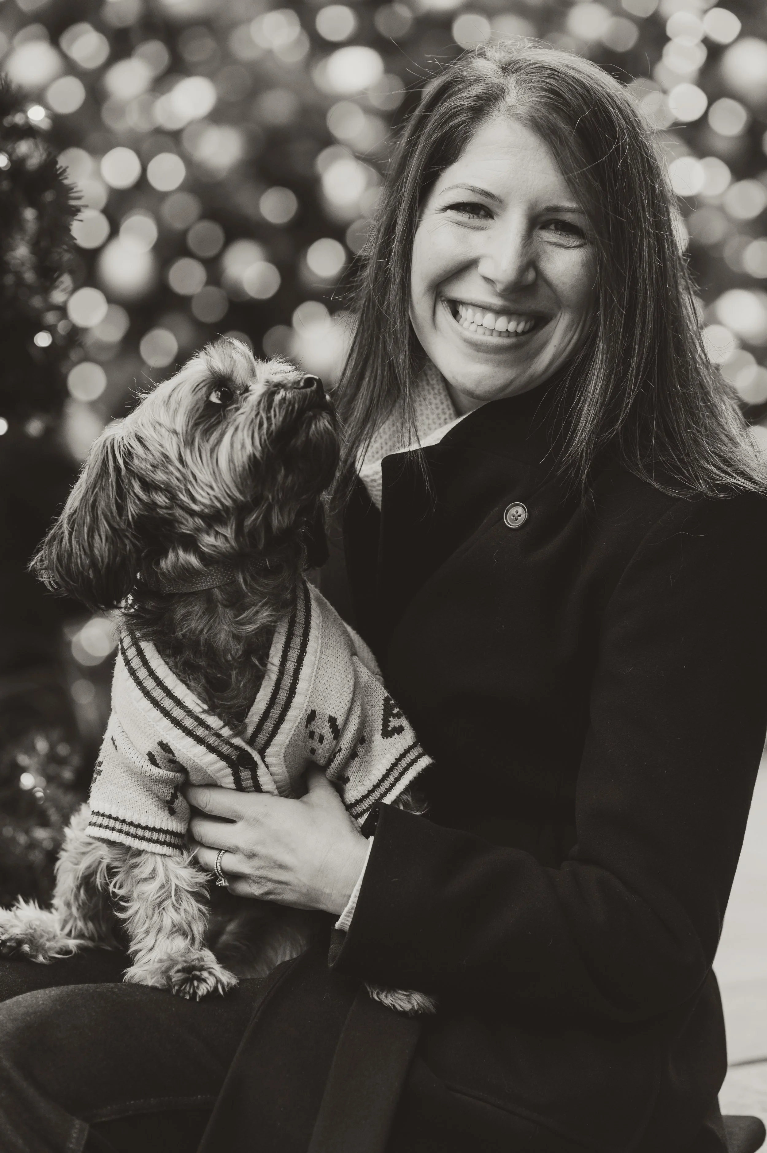 A woman smiling while holding a small dog in a Christmas sweater in front of a blurry decorated Christmas tree.
