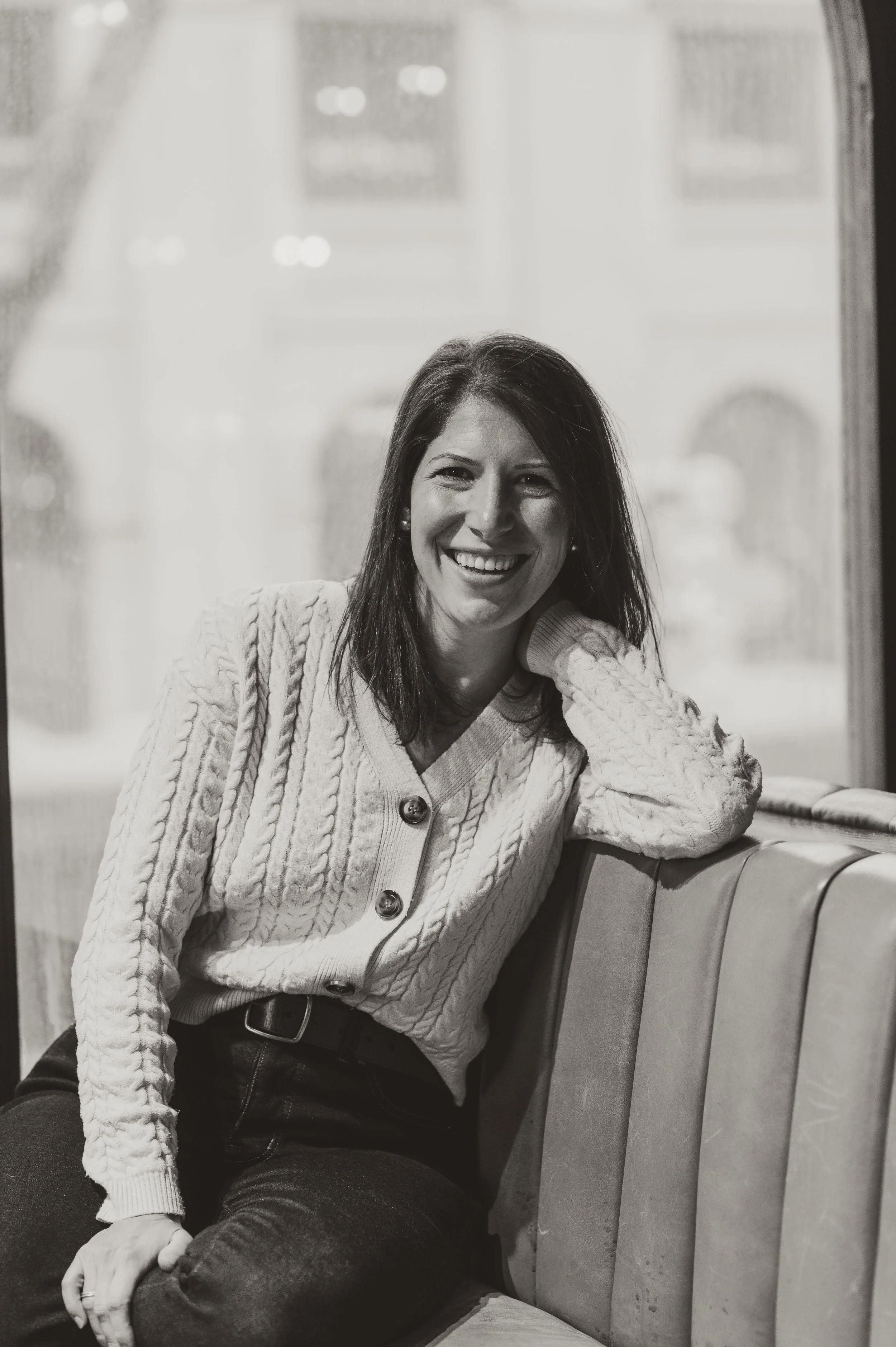 A woman in a knitted cardigan smiling and sitting on a bench by a window.