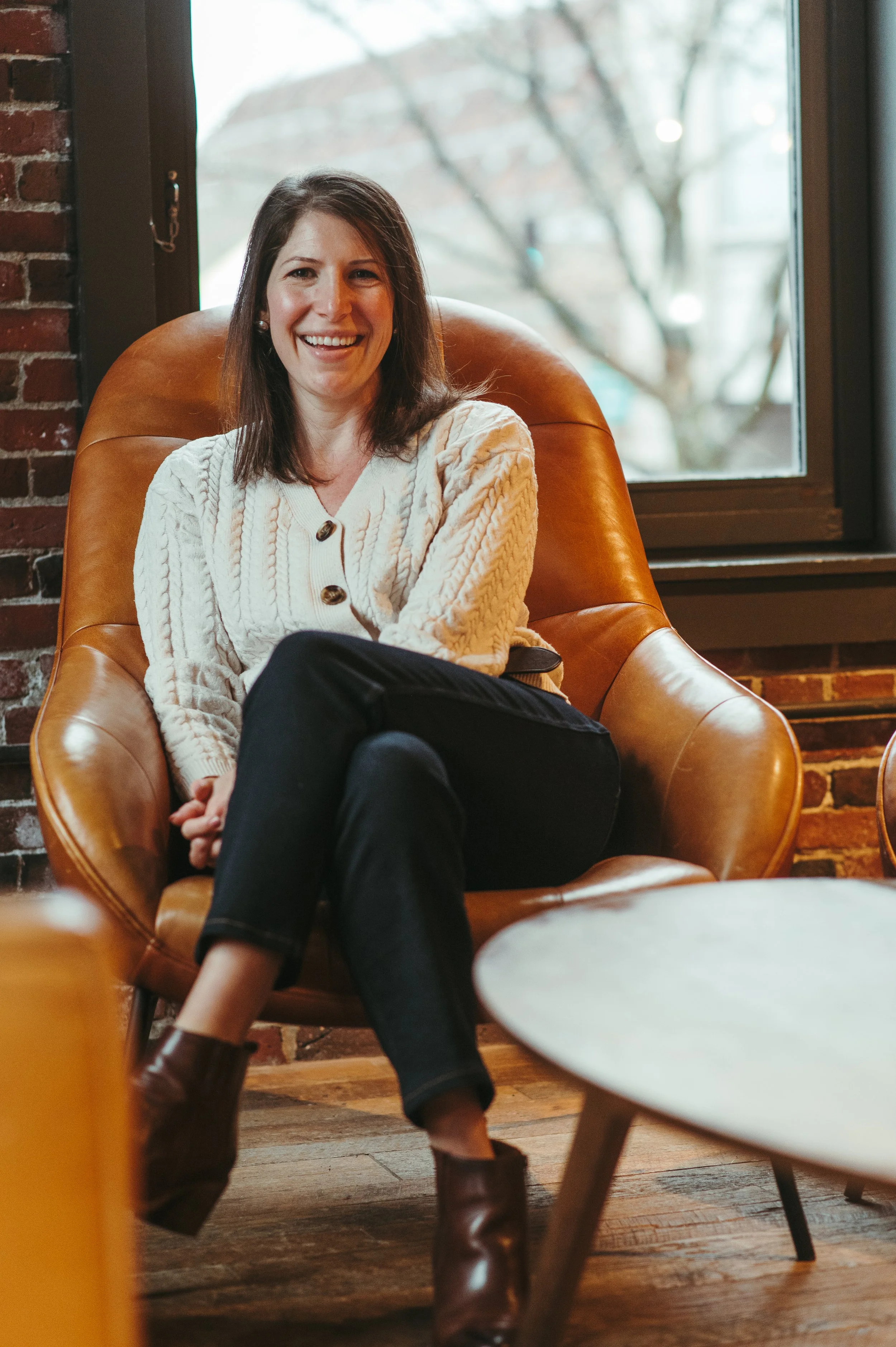 A woman with shoulder-length brown hair, smiling, sitting on a brown leather chair near a window, wearing a cream-colored cable-knit sweater, black pants, and brown ankle boots, in a cozy indoor setting.