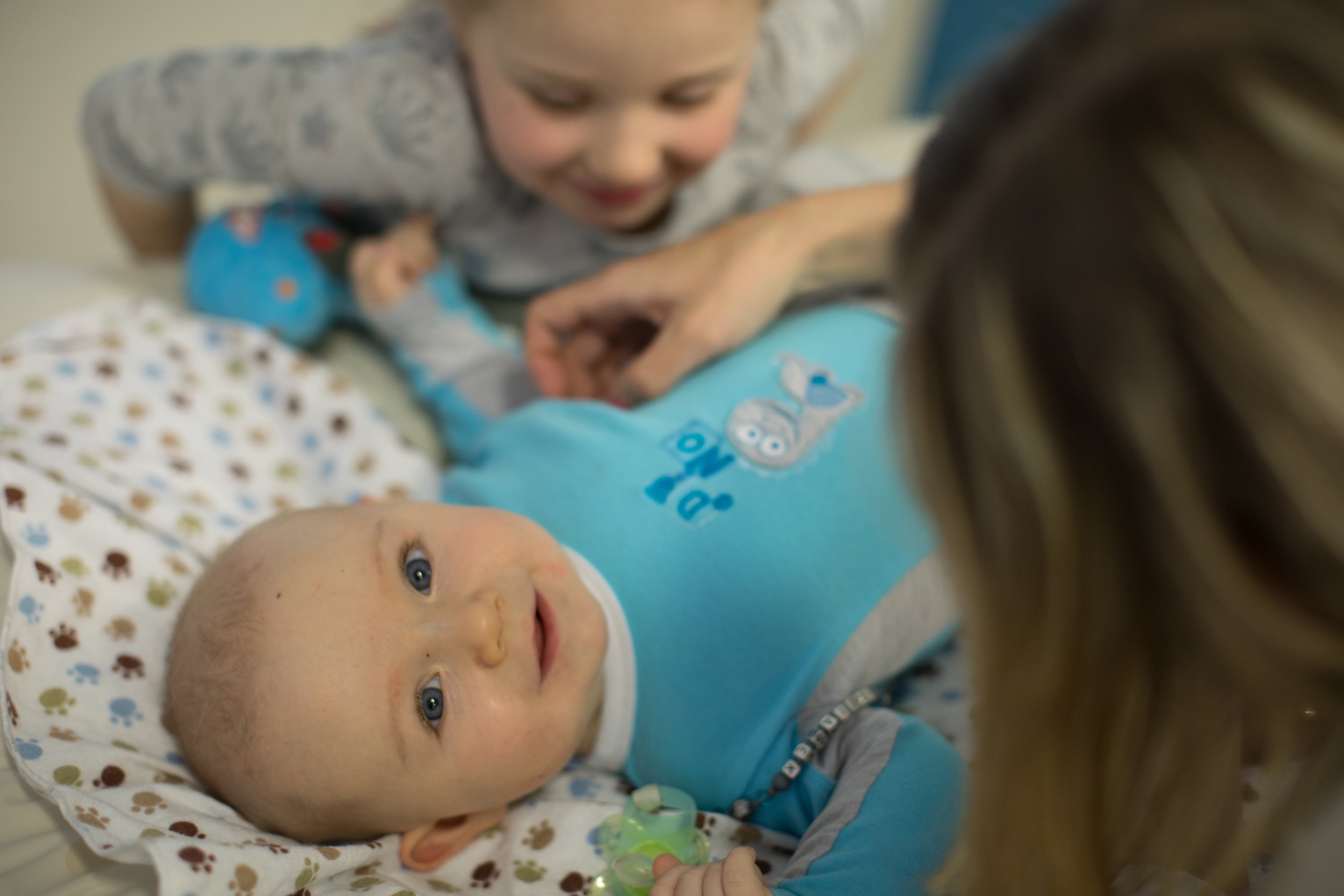 A smiling baby lying on a bed with a colorful blanket, looking up at an adult. The baby wears a blue shirt with a bear design. An adult with long brown hair is leaning over, holding or interacting with the baby. Another child, possibly a young girl, is in the background, also leaning towards the baby.