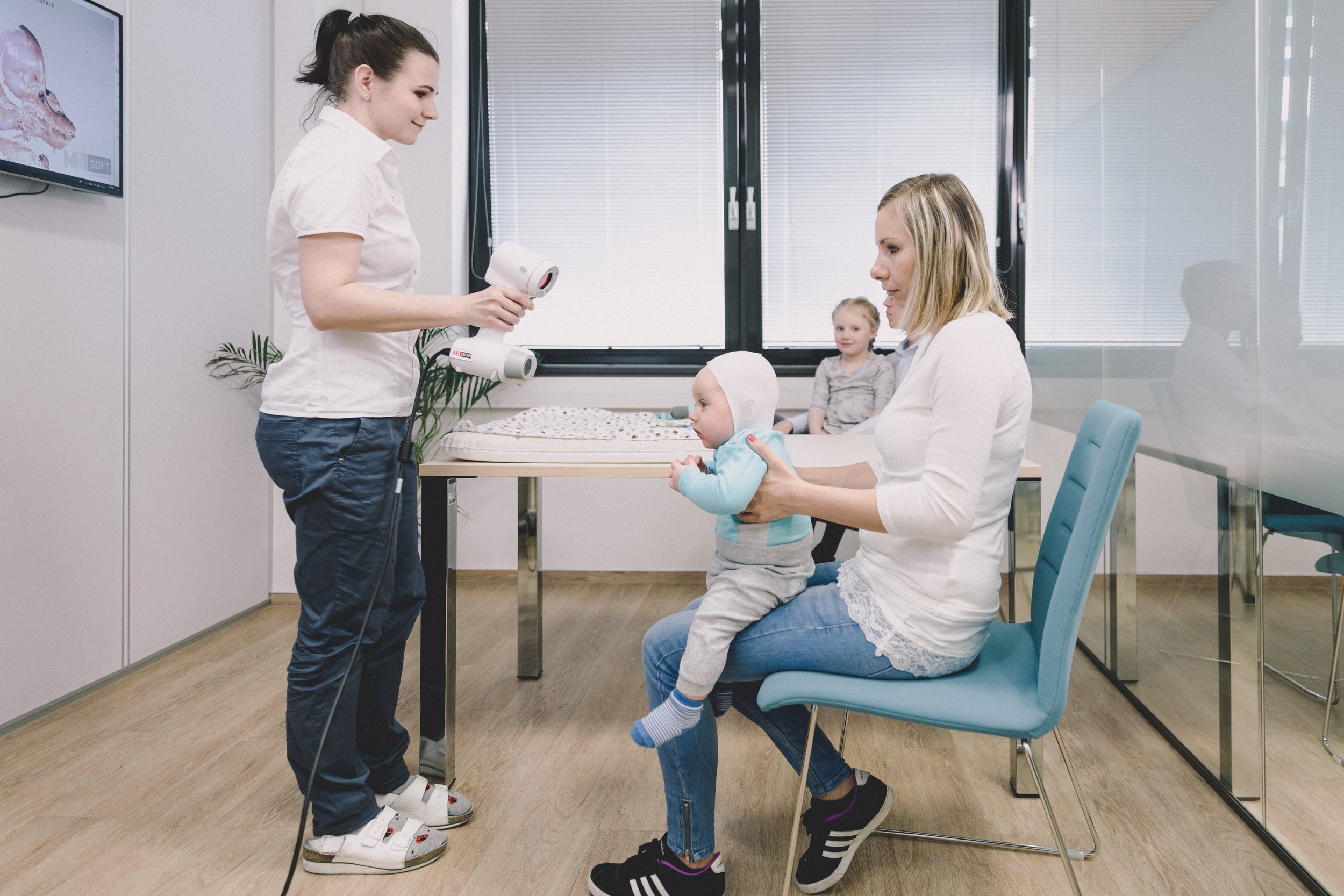 A mother holds a baby in a medical examination room while a nurse prepares to use a thermometer on the baby. In the background, another young girl is sitting on a table smiling.