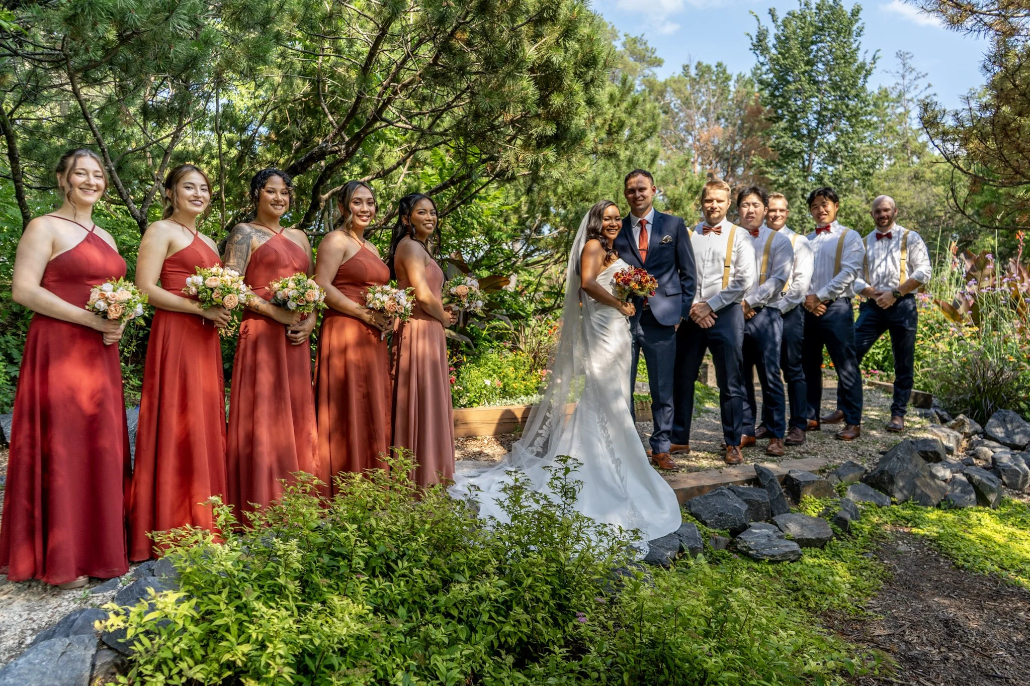 A wedding party outdoors on a sunny day, with nine women in red and pink dresses holding bouquets and two men, a bride in a white gown with a veil and a groom in a navy suit, standing together among green trees and plants.