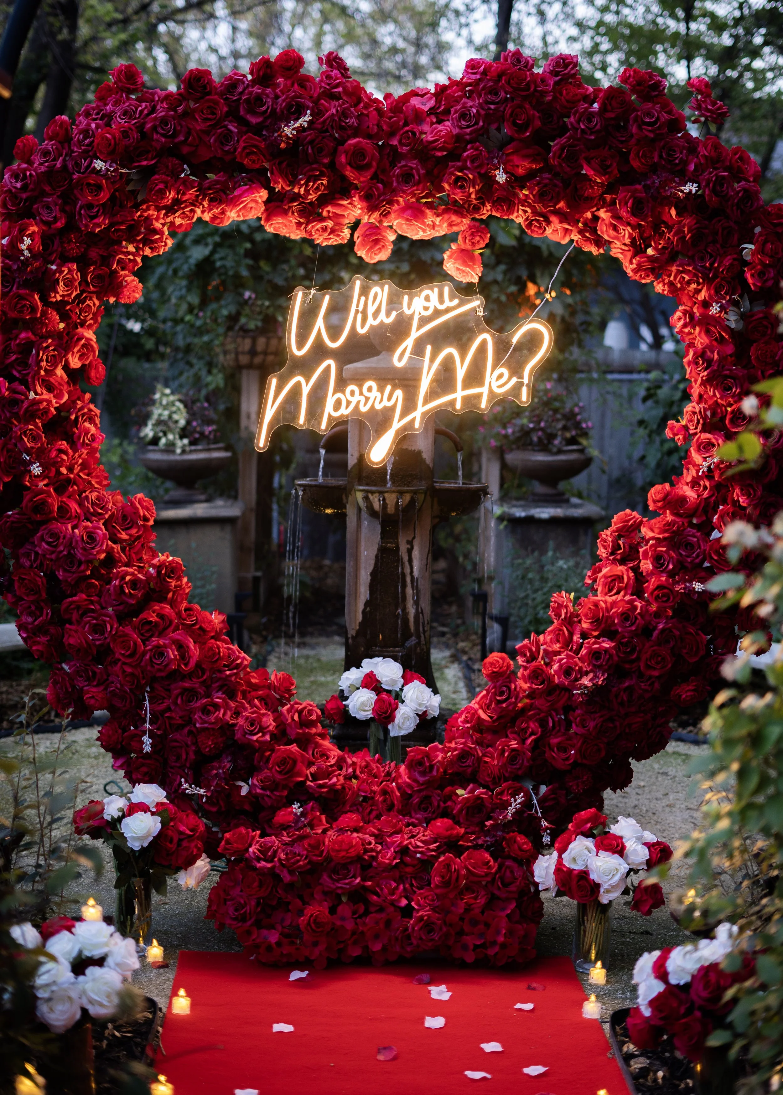 A romantic outdoor wedding setup with a heart-shaped floral arch made of red roses, illuminated neon sign asking 'Will you Marry Me?', a red carpet aisle with white and red rose petals, and small candles along the sides.