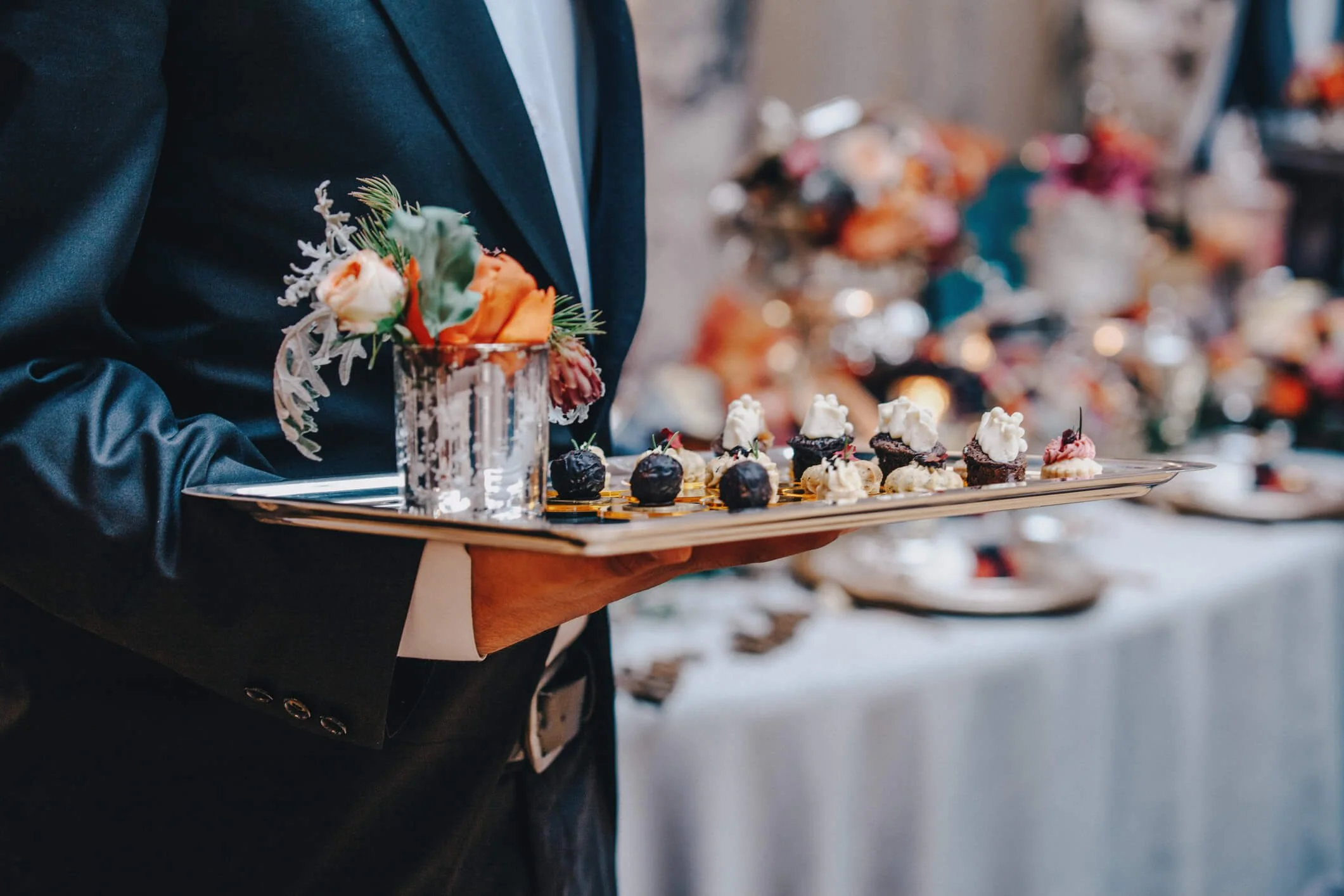 Person in formal attire holding a tray of decorated desserts and a floral arrangement in a glass vase at an event or party.