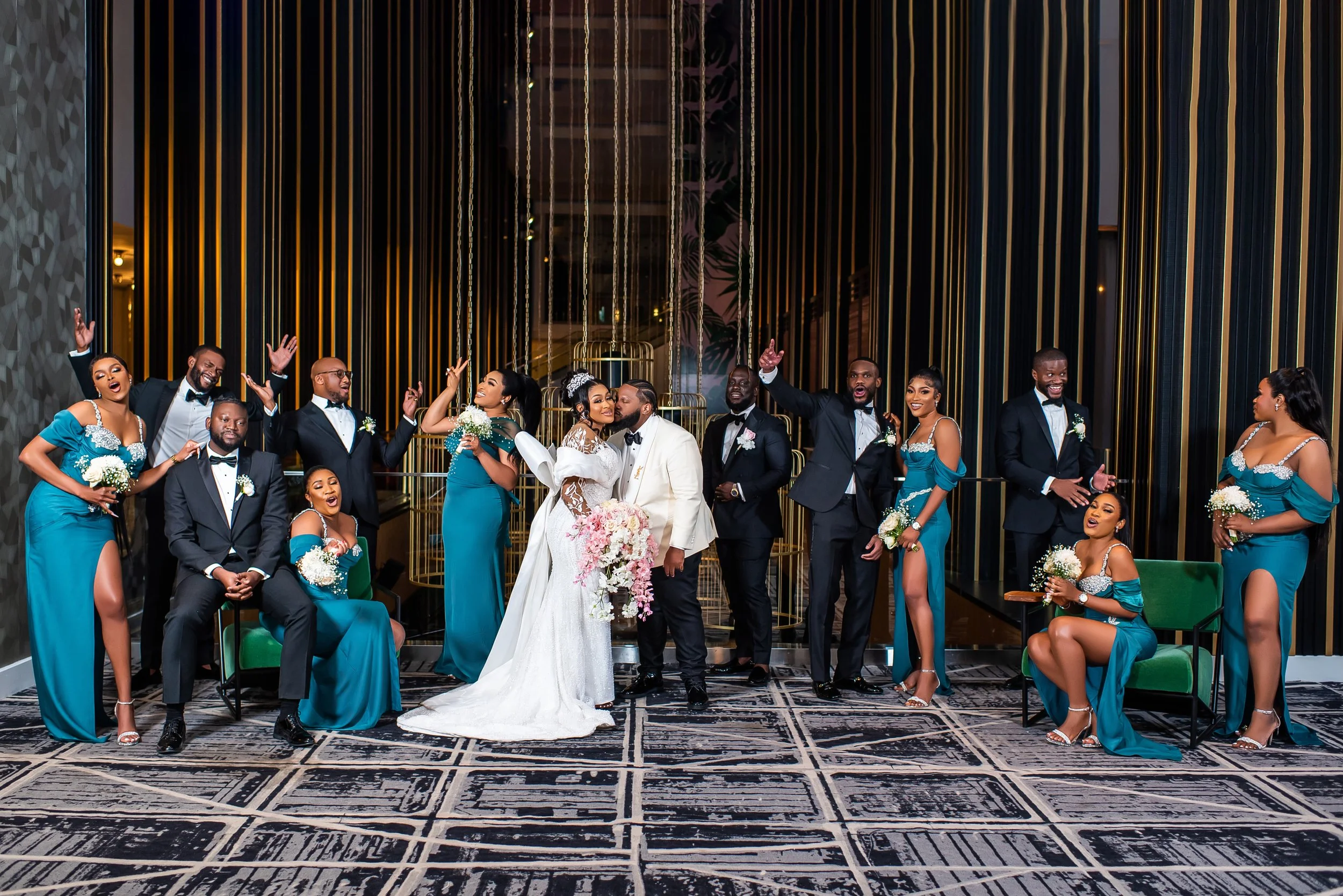 A wedding party standing and sitting in a modern, elegant venue with black and gold decor. The bride and groom are kissing in the center, surrounded by bridesmaids and groomsmen dressed in tuxedos and teal dresses with floral arrangements. Everyone is smiling and celebrating.