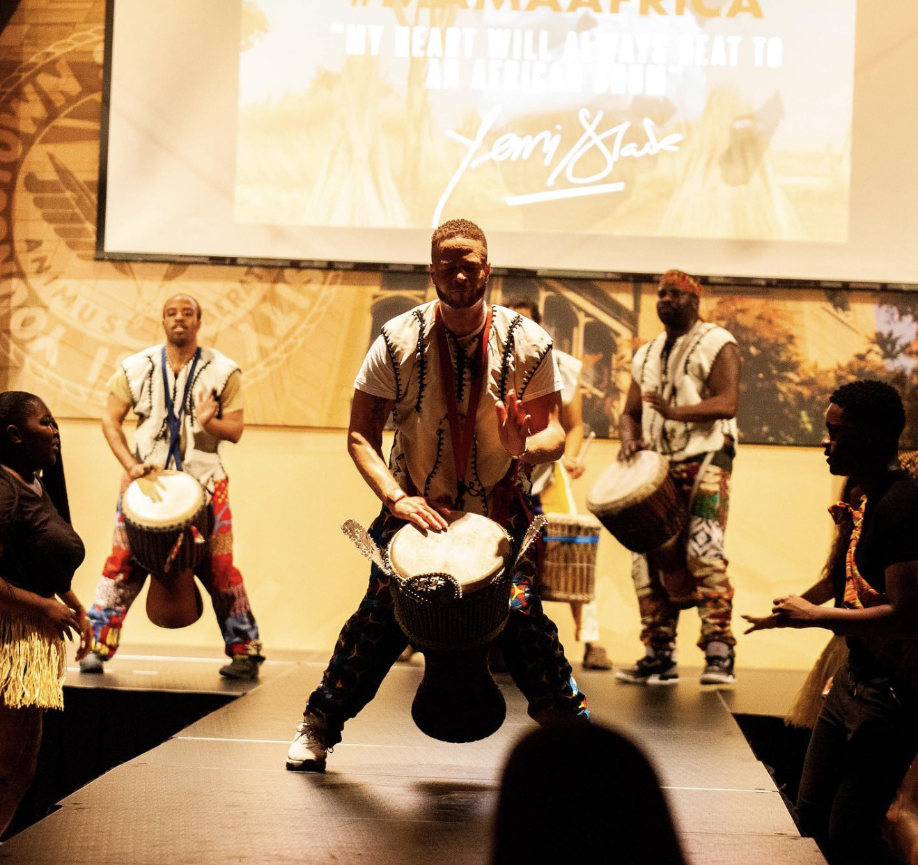 A group of people performing a traditional African dance on stage, with some playing drums and others dancing. The background features a large screen with text and a signature.