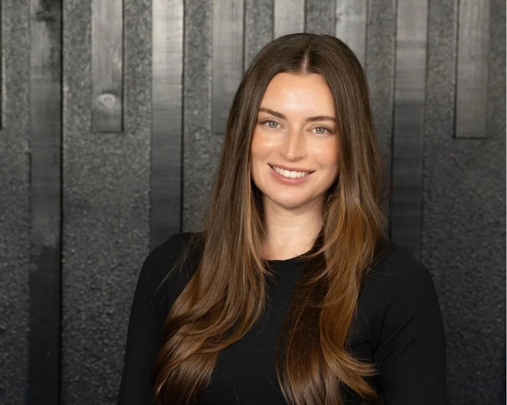 A woman with long brown hair, smiling, wearing a black top, standing against a dark textured wooden wall background.