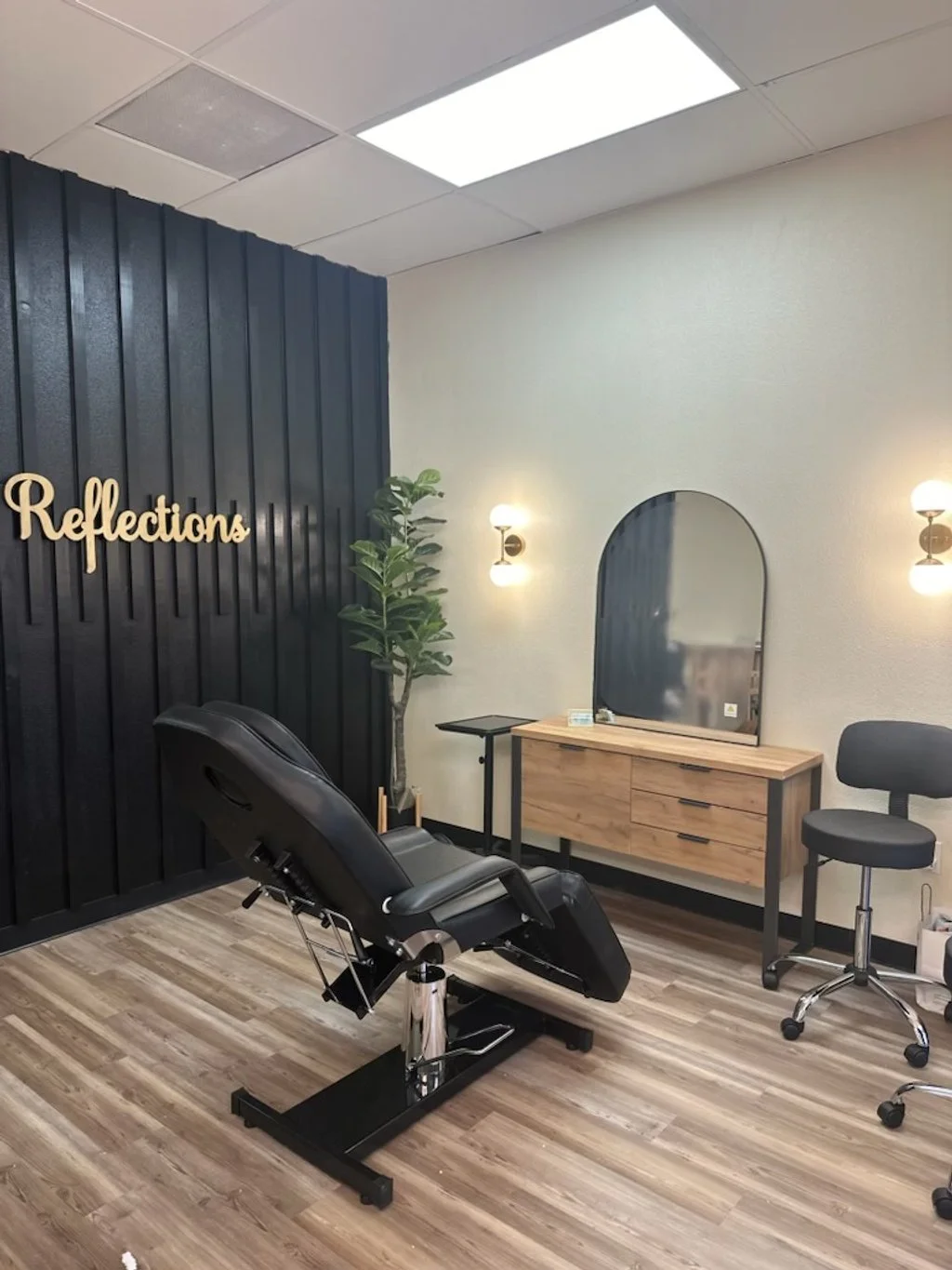 A modern beauty or spa treatment room with a black adjustable reclining chair, a large oval mirror on a wooden dresser, a small black side table, a black rolling stool, and a tall green plant. The wall behind the chair has a black panel with the word "Reflections" in cursive. Two wall-mounted lights illuminate the room, which has a wood-patterned floor and a ceiling with fluorescent lighting panels.