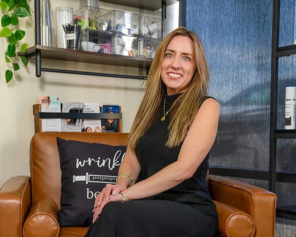 A woman with long blonde hair sitting on a brown leather couch in an office or clinic setting, smiling at the camera. She's wearing a black sleeveless dress and gold jewelry, with a pillow that says 'wrinkle be gone' on her right side. Behind her are shelves with books, magazines, and office supplies, and a plant on the left side.