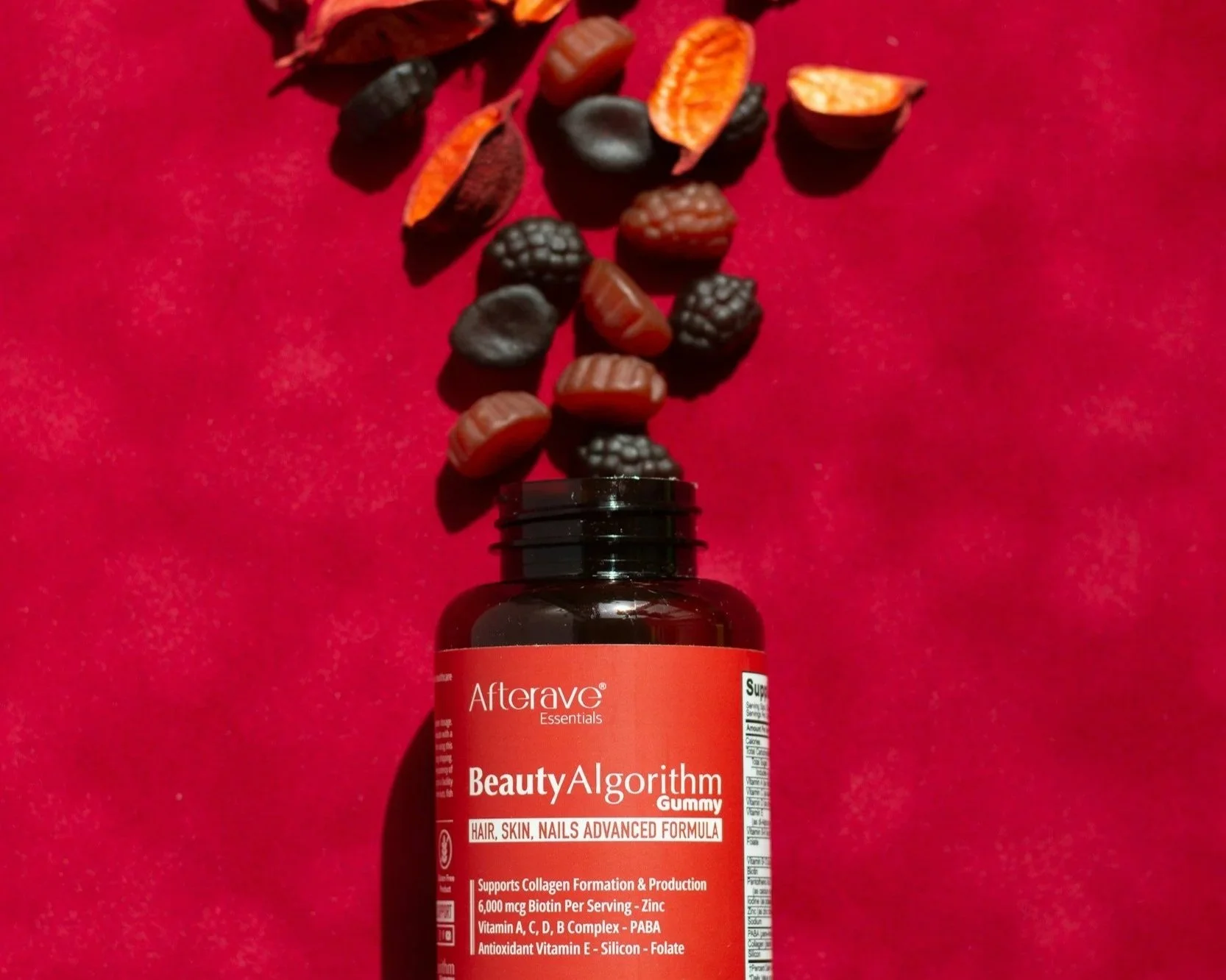 A red bottle of Afterave Essentials BeautyAlgorithm Gummy supplement with black and red gummy supplements spilling out onto a red surface.