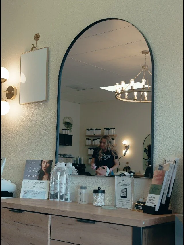 A mirror reflecting a woman in a hair salon, with shelves of hair products and salon equipment in the background, and bottles of hair care products on a wooden counter in the foreground.
