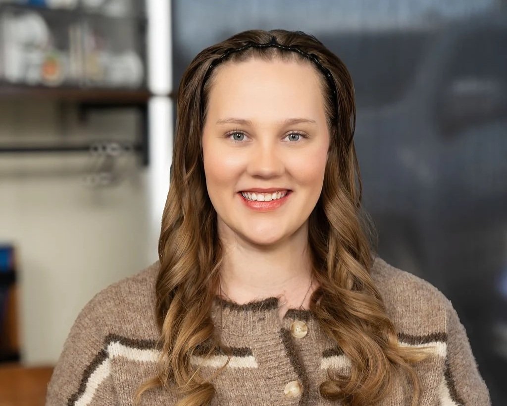 A young woman with long, wavy brown hair and a headband, smiling at the camera, wearing a beige sweater with a brown and white pattern, indoors with a blurred background.