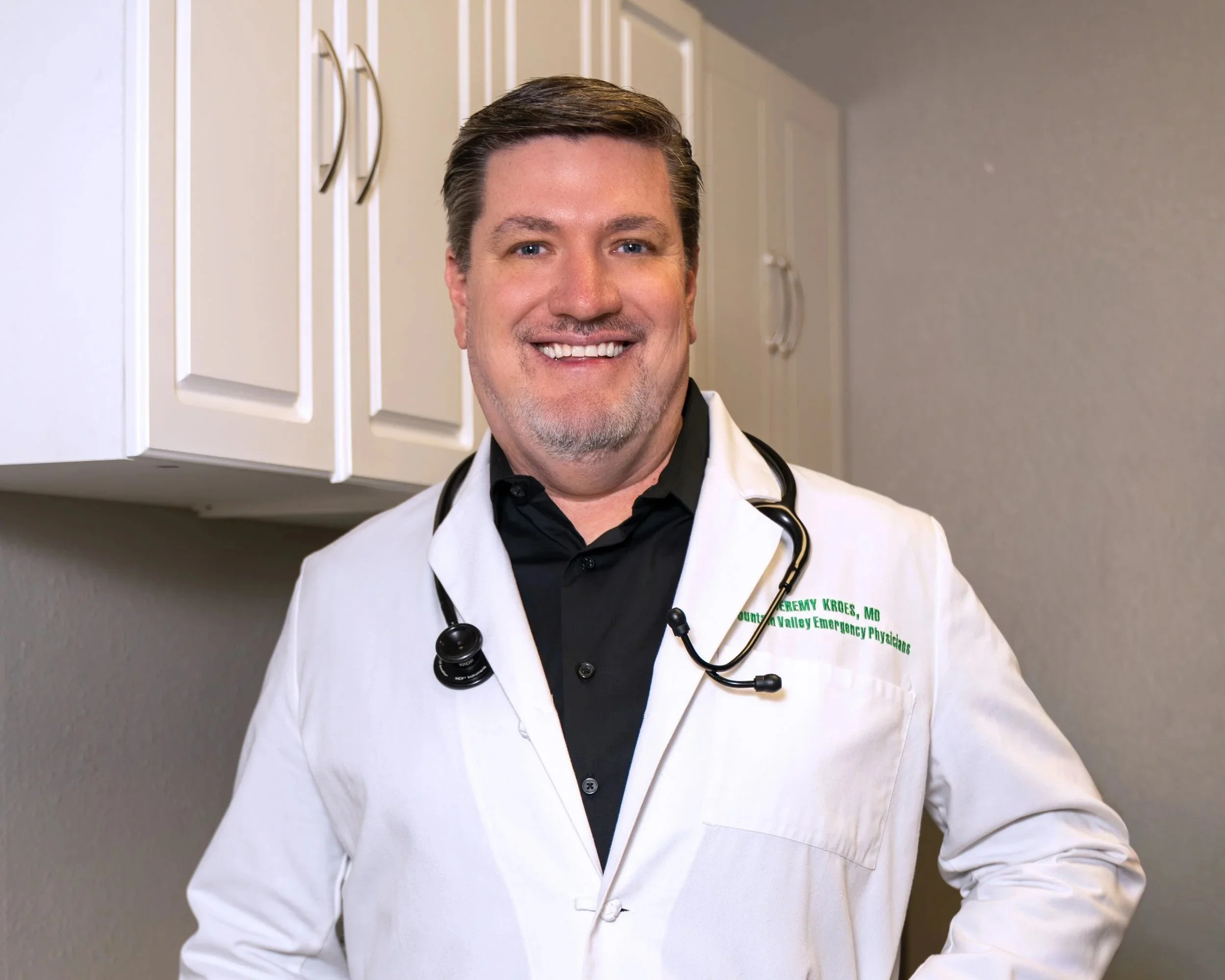 A smiling male doctor wearing a white lab coat with green embroidered text, a black shirt, and a stethoscope around his neck, standing in a medical office with white cabinets in the background.