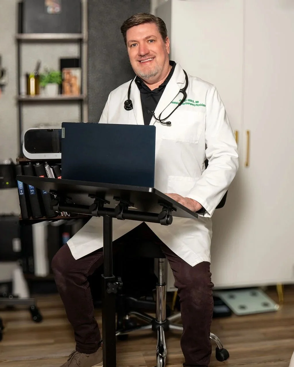 A male doctor or medical professional in a white coat with a stethoscope around his neck, sitting at a desk with a laptop in a medical or office setting.
