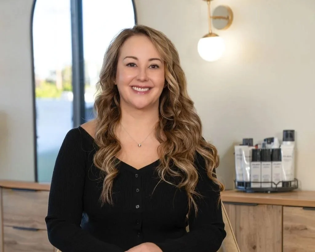 A smiling woman with long, curly hair sitting indoors near a window, with a wooden counter and skincare products in the background.