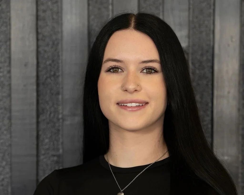 A young woman with long black hair, light skin, and a nose ring, wearing a black top and a silver necklace, smiling at the camera against a dark textured wooden background.