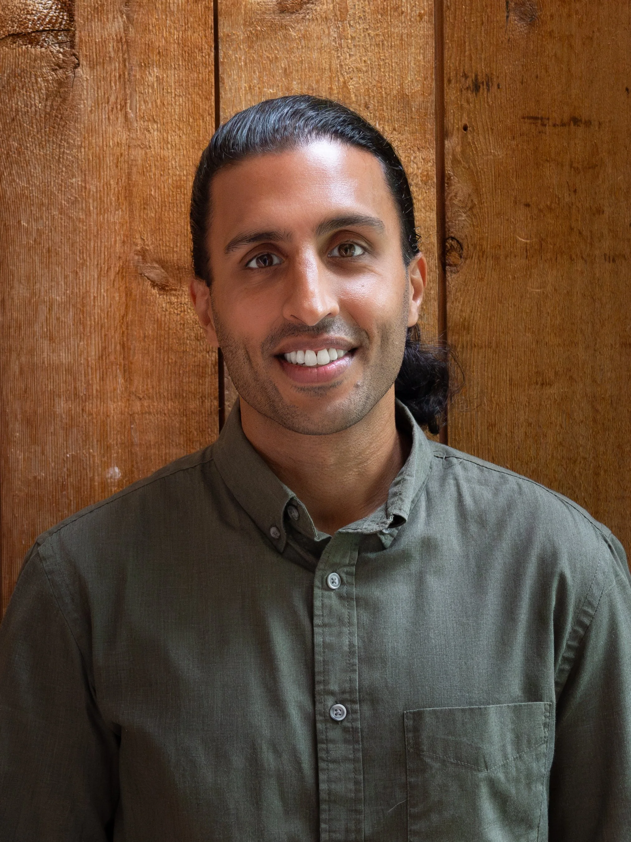 A man with dark hair tied back, wearing an olive green button-up shirt, smiling in front of a wooden background.