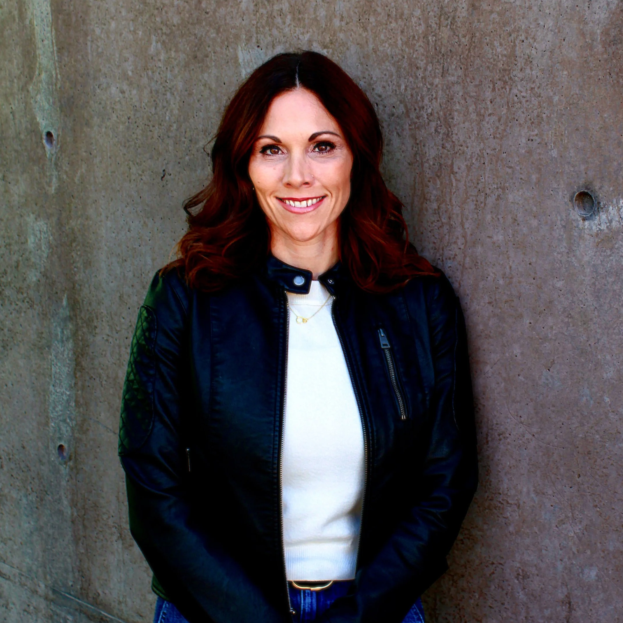 A woman with shoulder-length dark red hair, smiling, standing against a textured concrete wall, wearing a black leather jacket over a white top.